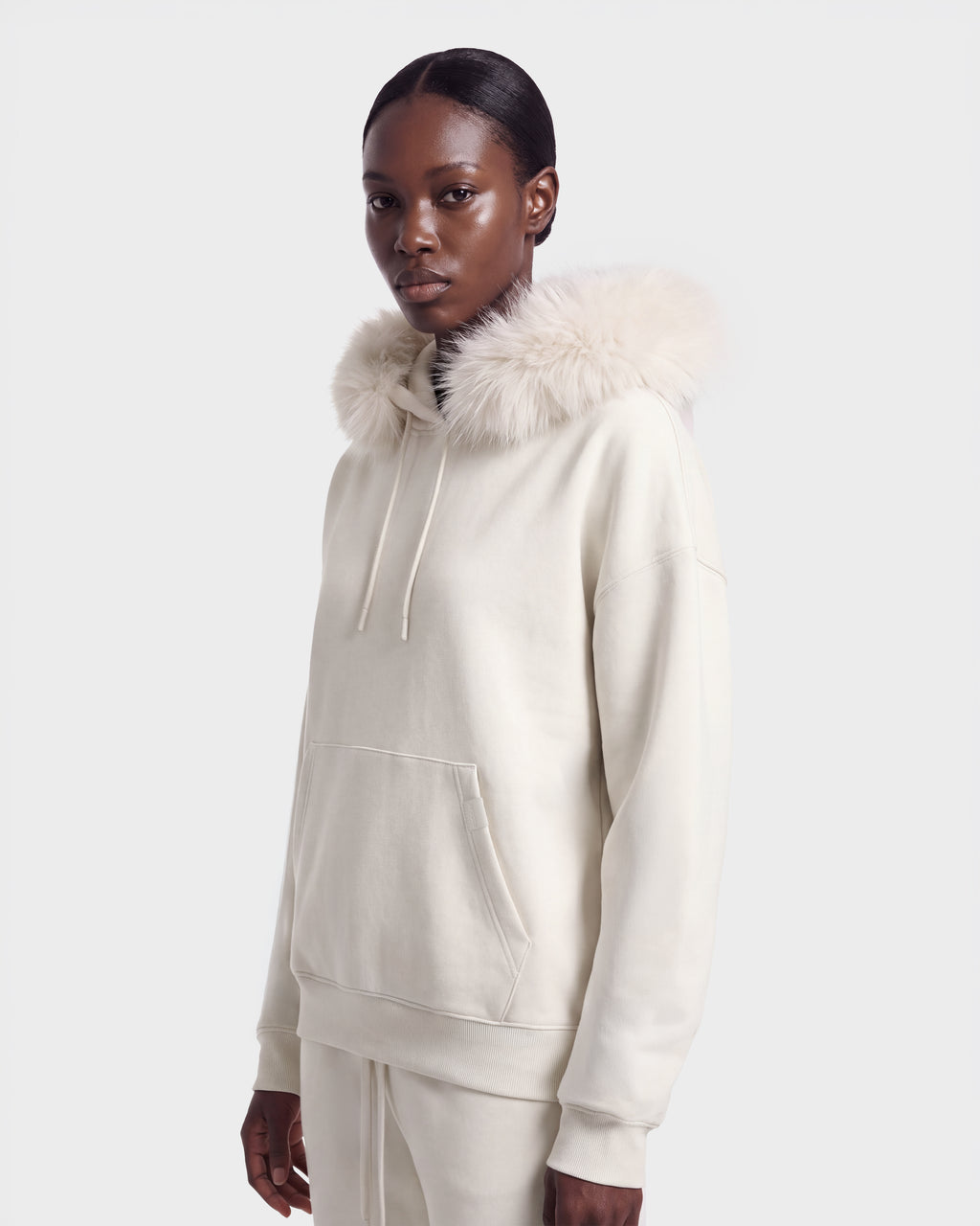 A person wearing the MILENA - 8526689 Vanilla hoodie by Rudsak, featuring a cream color and fluffy faux fur hood trim, stands against a plain, light background.