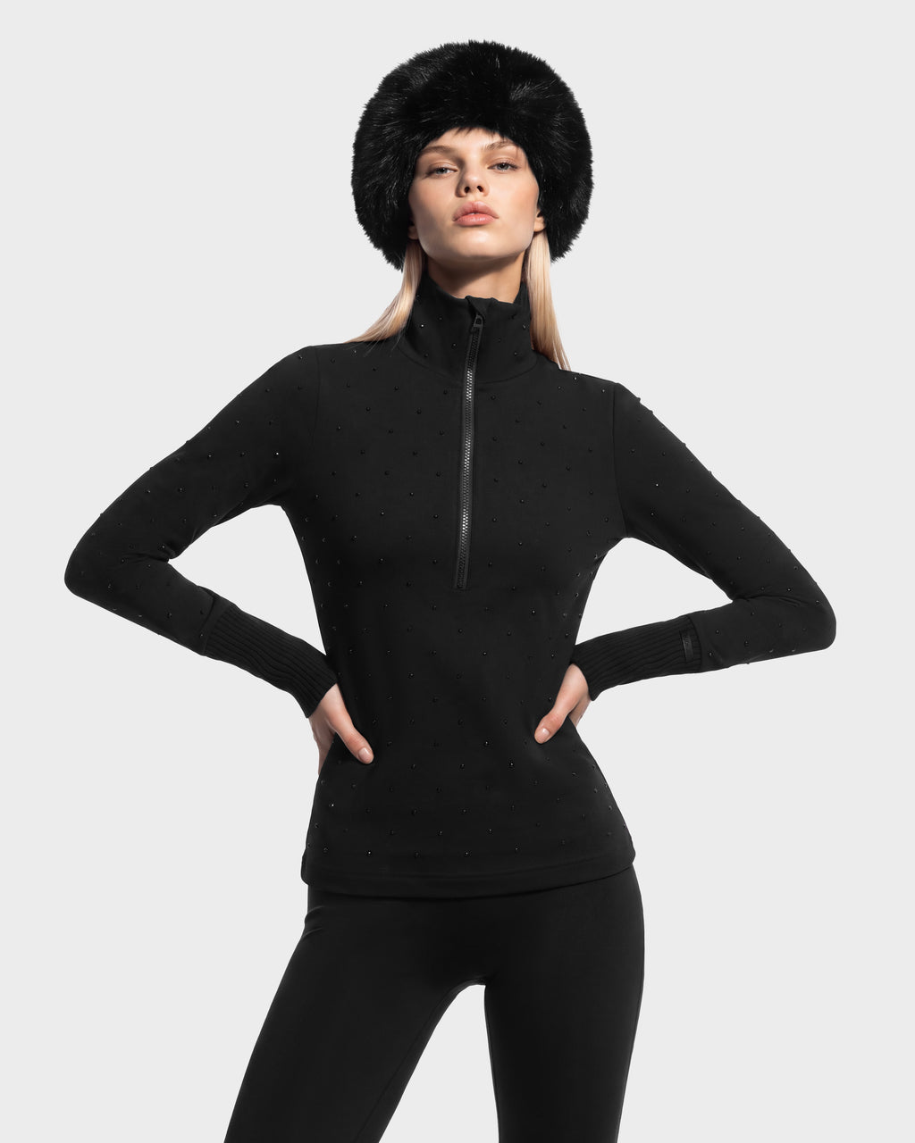 A person wearing a black Rudsak NEVA - 8526524 Black long-sleeve half-zip top with studded detailing, black leggings, and a black faux fur hat.