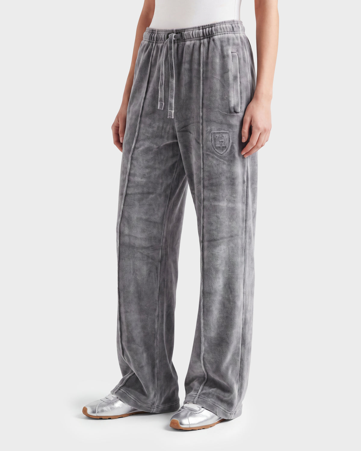 A person wears the Rudsak OLIVYA HV - H526903 Titanium gray velour sweatpants featuring a drawstring waist, side pockets, and an embroidered crest on the left thigh, styled with a white top and silver sneakers.