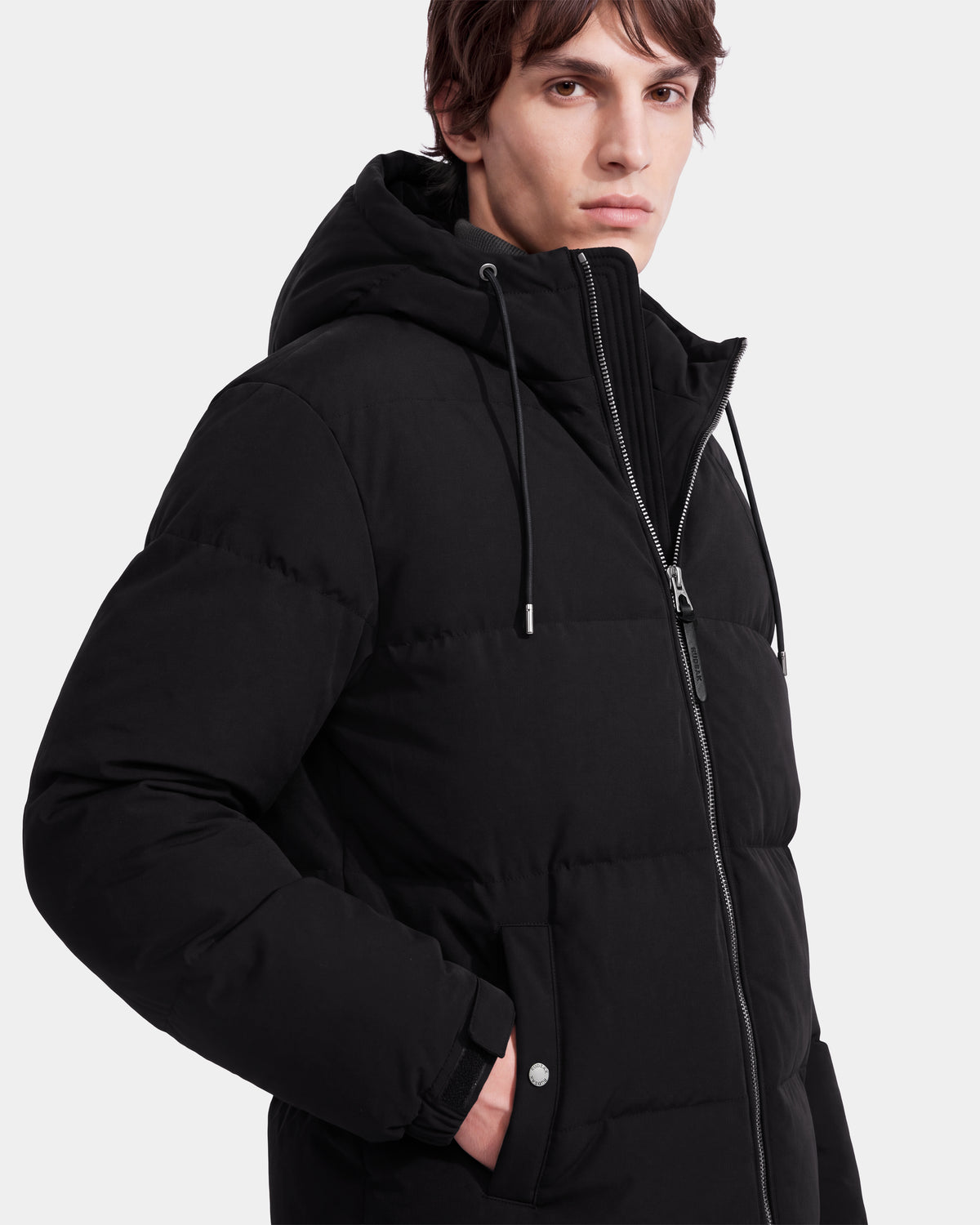 A person with short brown hair wears the Rudsak STERLING EL - 6126759 Black puffer jacket, featuring a hood, zipper, and snap buttons, while standing against a plain white background.