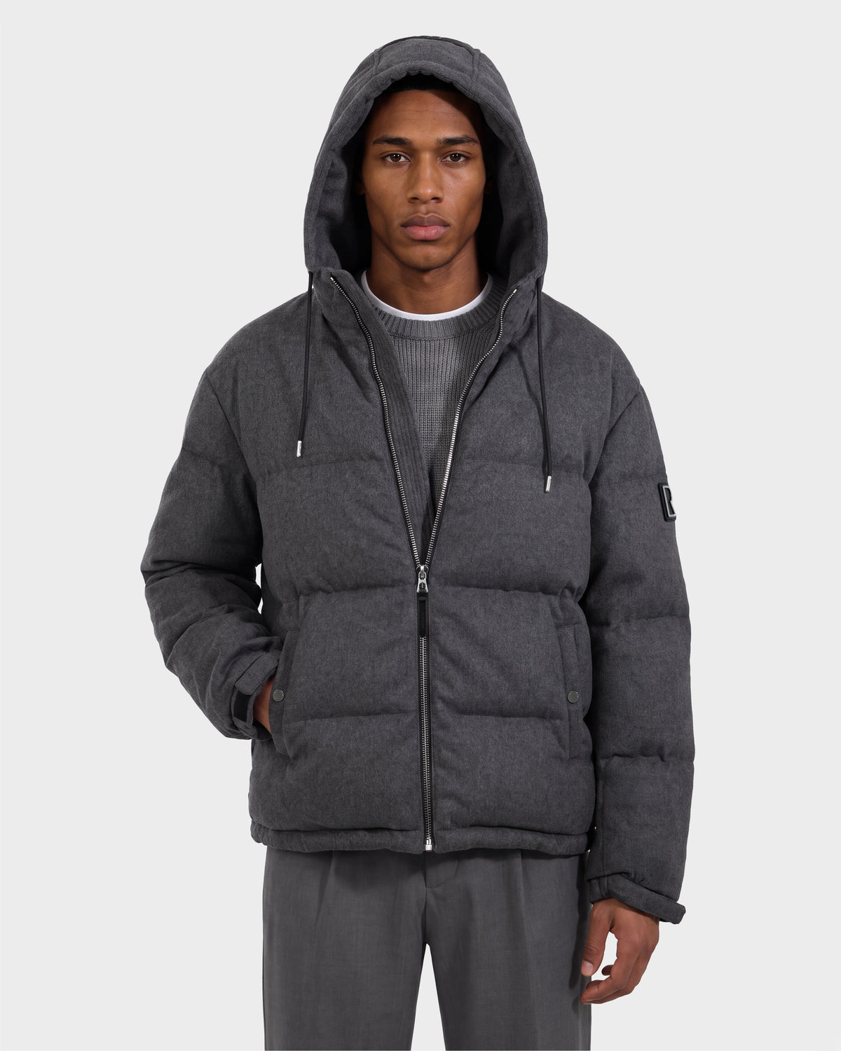 A person is seen wearing the Rudsak STERLING W - 6126760 Heather Charcoal puffer jacket with a hood, paired with gray pants and a ribbed gray sweater. The jacket is zipped up, and the individual has one hand tucked into a pocket, standing against a plain light background.