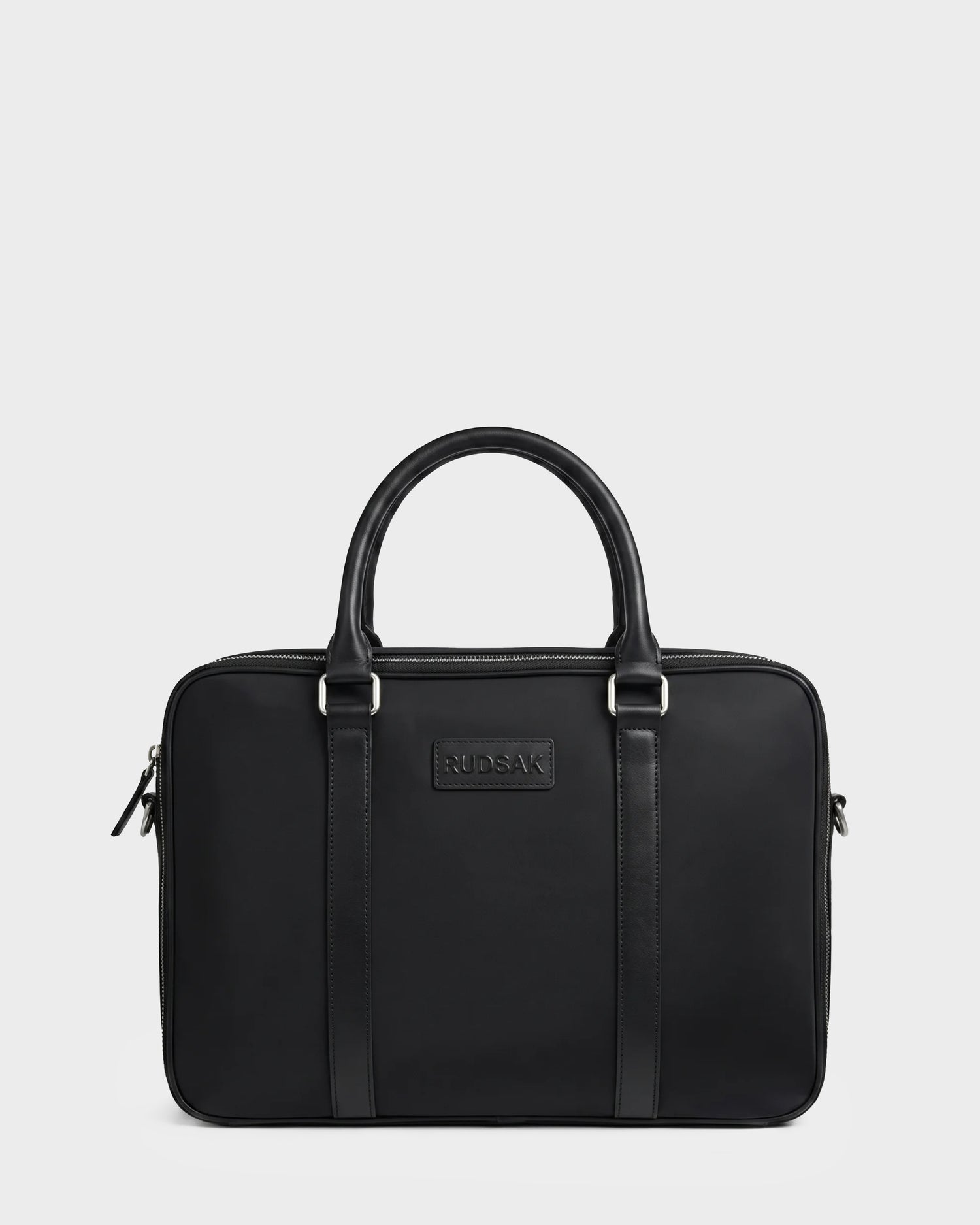 The HENRY N - 9326056 Black by Rudsak is a rectangular black leather briefcase featuring two handles, silver hardware, and the Rudsak logo embossed at the front center. The bag is displayed against a plain white background.
