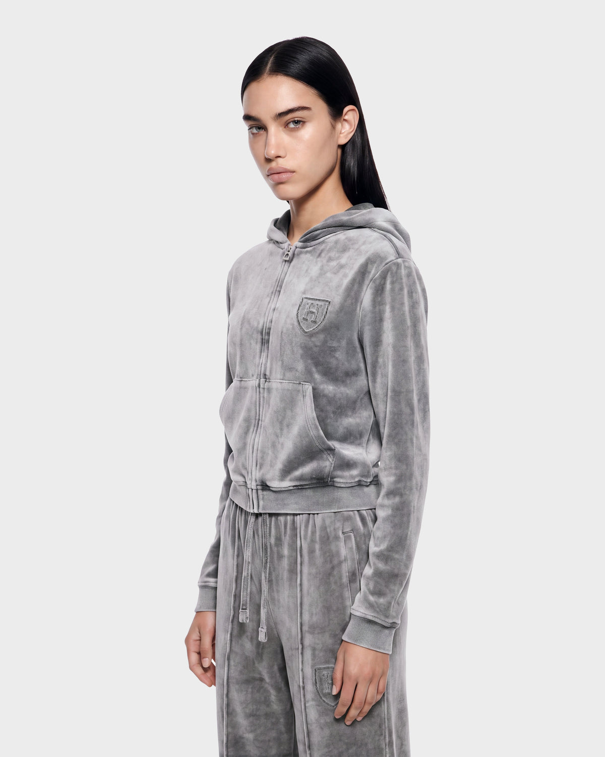 A person with long dark hair wears the Rudsak TYRA HV - H526904 Titanium, a light gray velour tracksuit with a hood, zip-up front, and pockets, standing against a plain light background.