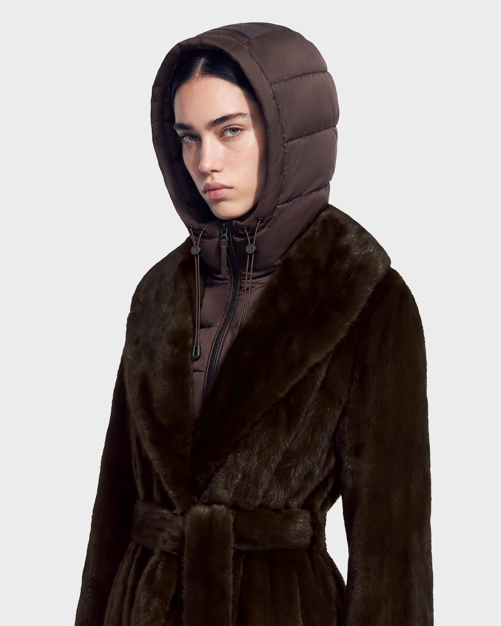 A person wears a dark brown hooded Rudsak VIVIENNE - 8126528 Espresso duck down jacket layered under a belted faux fur coat for winter city dressing.