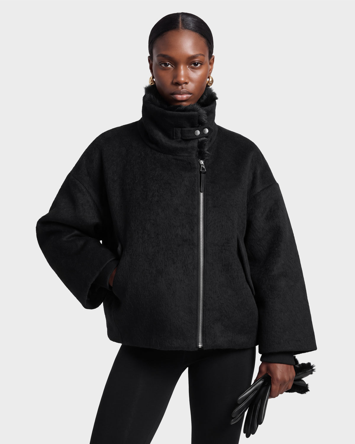 A woman models the Rudsak ZARIA - 8126999 Black high-collar jacket and matching leggings, holding black gloves. She wears gold earrings and maintains a serious expression while posing against a plain light gray background.