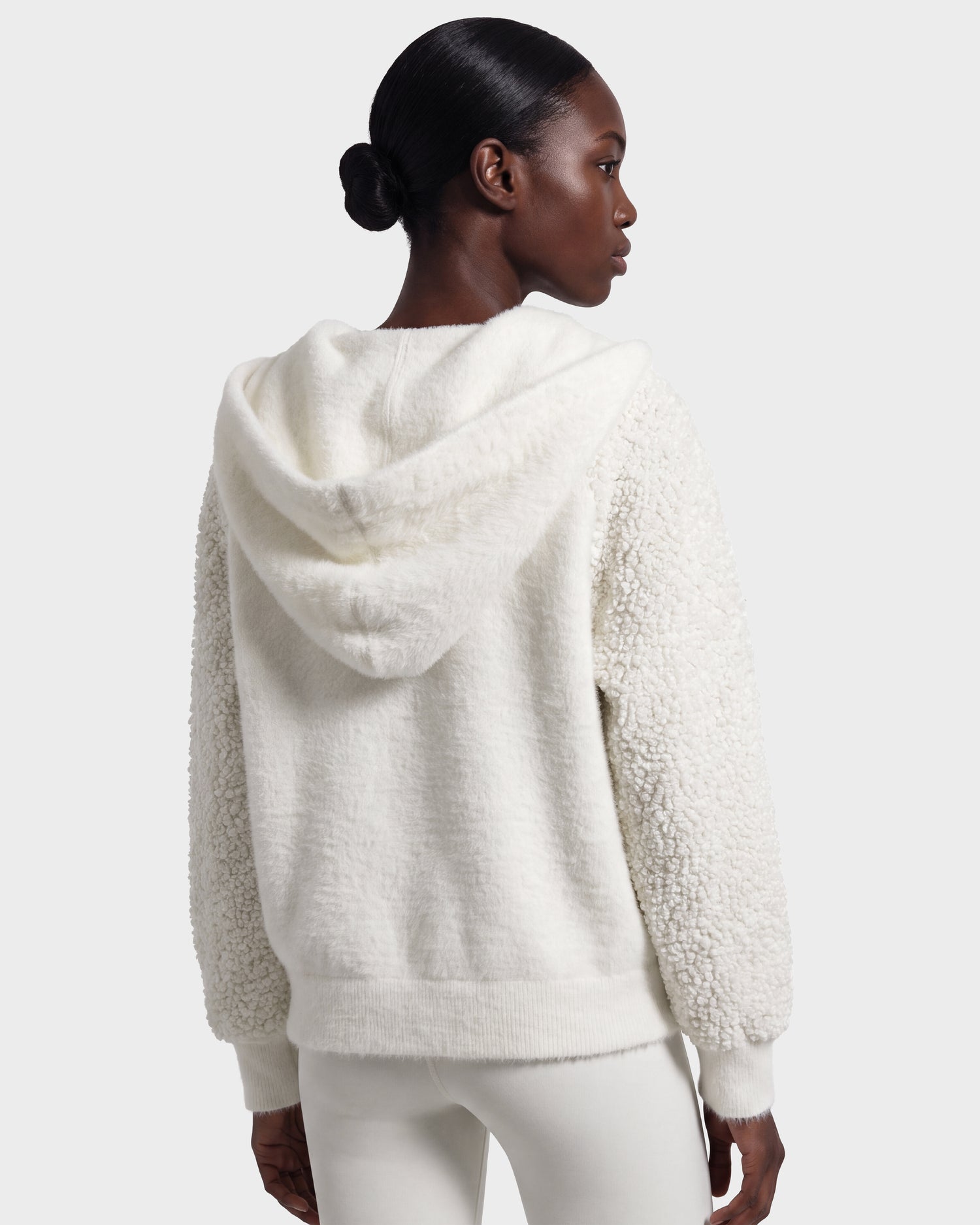A woman with dark hair in a low bun is shown from behind against a plain light gray background, wearing the Rudsak DAKOTA - 8526515 Iceland textured white hooded jacket and white pants.
