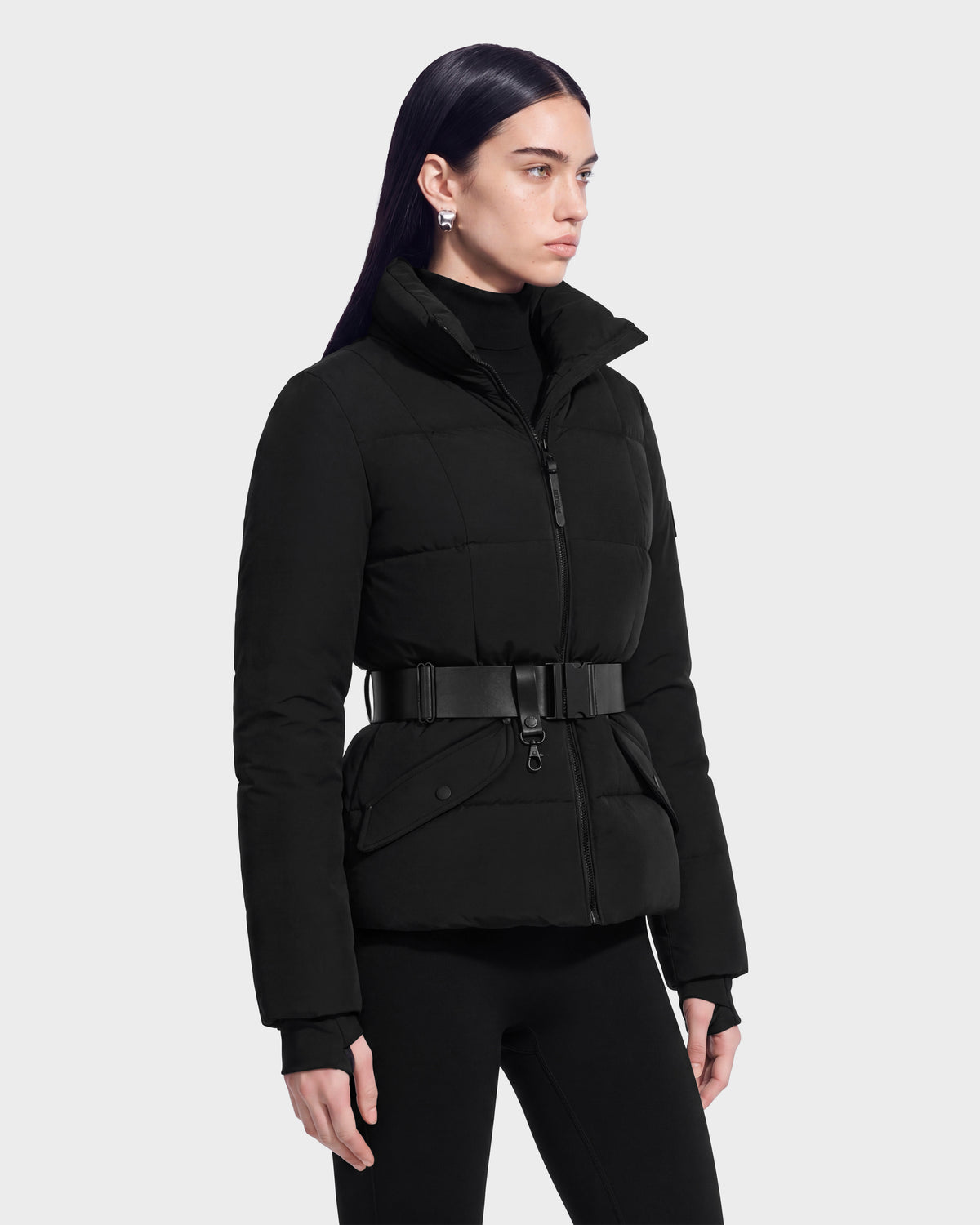 A woman stands against a plain light background, looking to the side with a neutral expression while wearing the Rudsak ADELINE - 8124544 Black belted quilted short jacket over a black turtleneck and black pants.