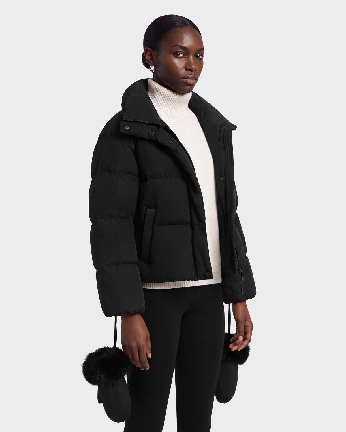 A person wears a black Rudsak ALONDRA - 8126992 Black puffer jacket over a white turtleneck, with black mittens hanging from the sleeves.