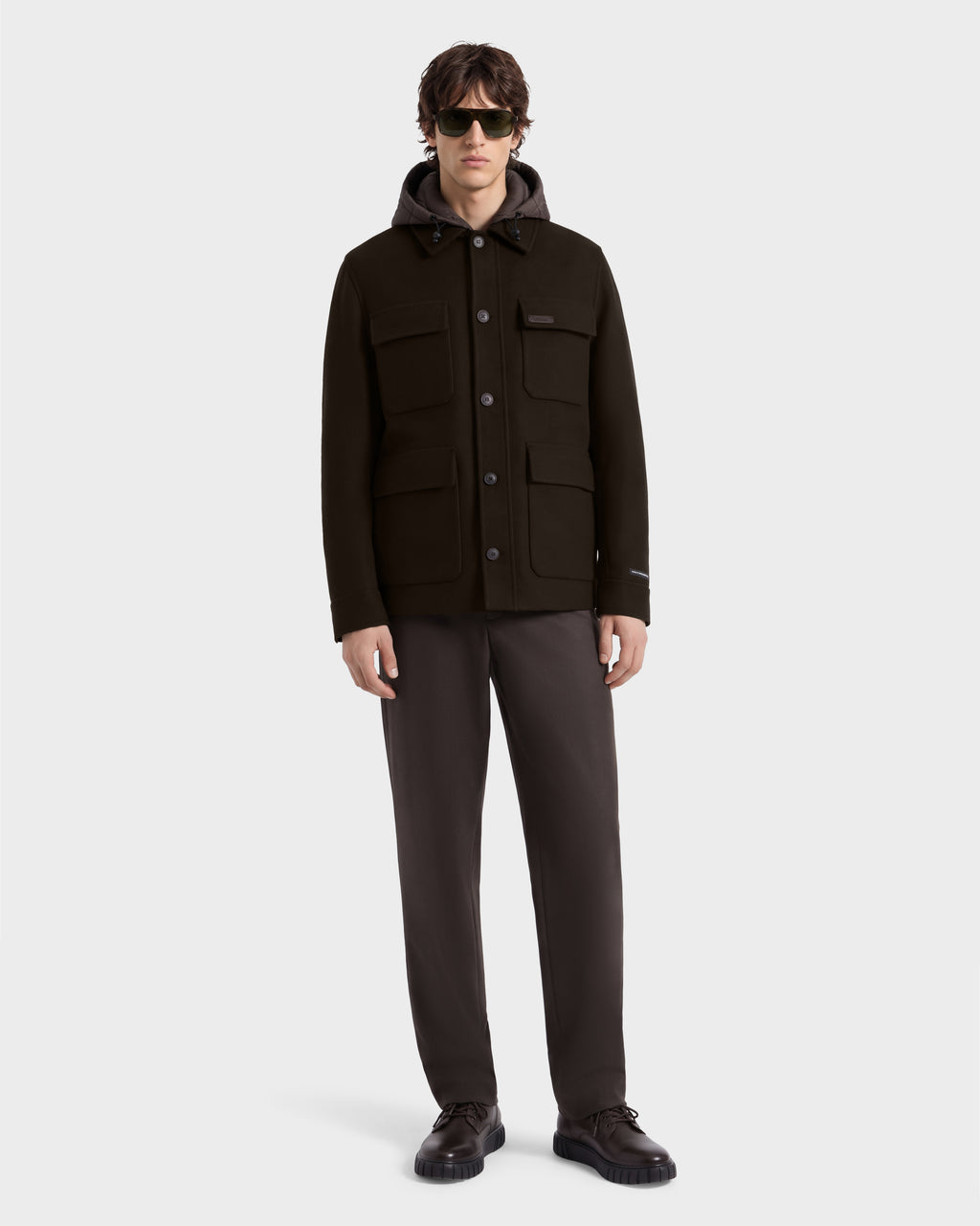 A man wearing a BRUNO - 6126716 Espresso jacket with a hood, dark trousers, dark sunglasses, and dark shoes.