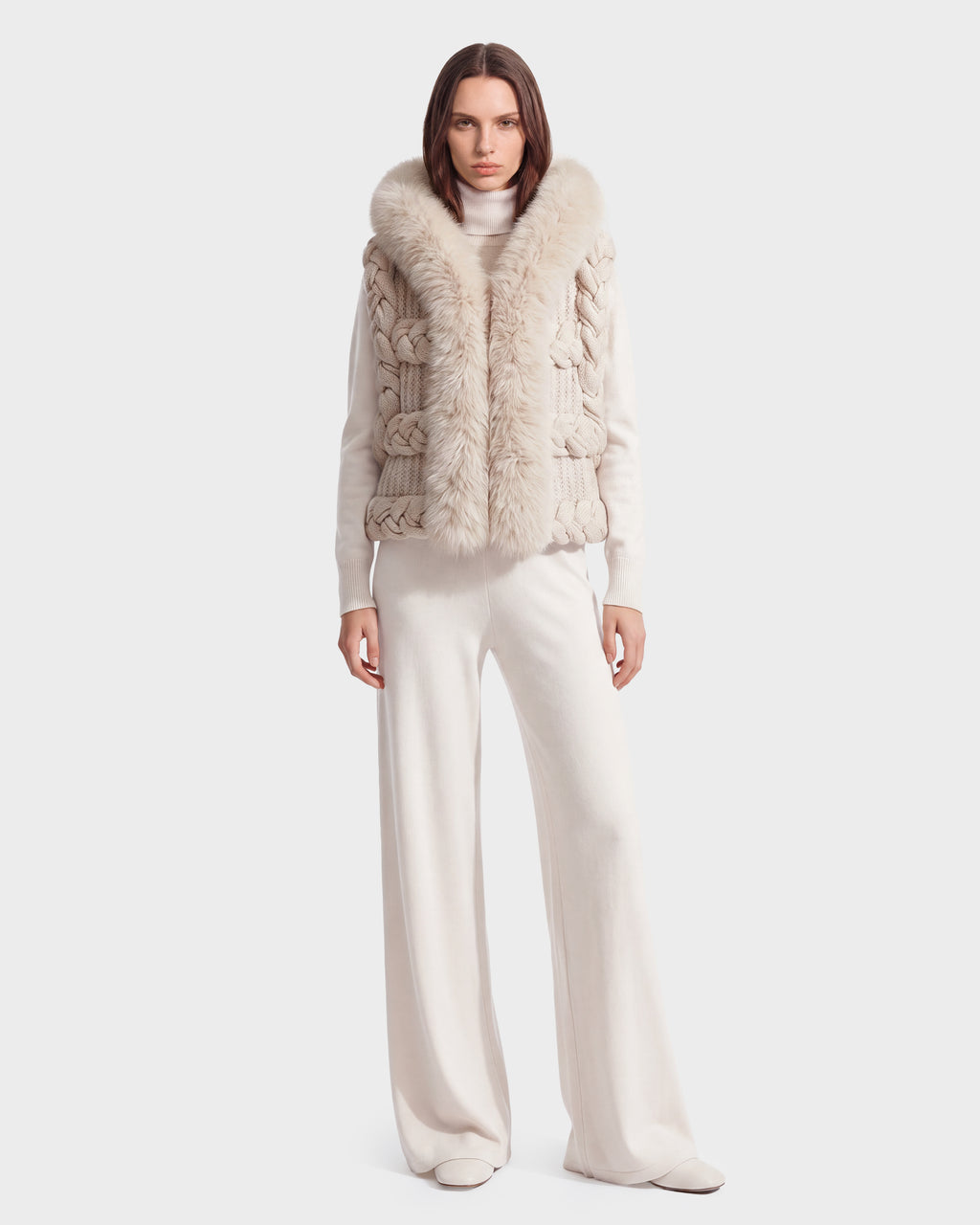 A woman stands facing forward against a plain white background, effortlessly capturing the essence of winter layering in the CICELY - 8526686 Vanilla textured beige knit vest by Rudsak, styled over a white turtleneck and wide-leg pants.