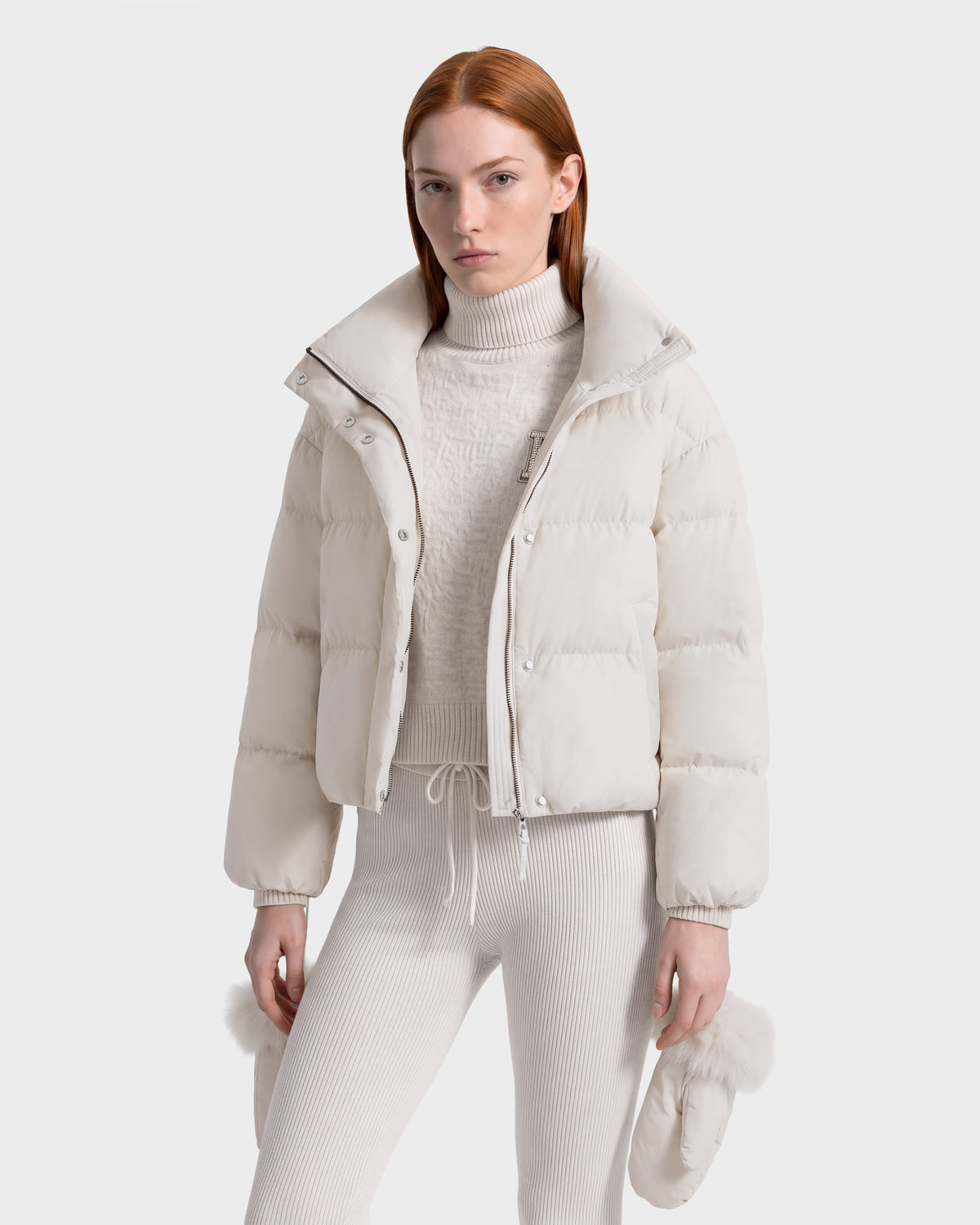 A woman wearing a Rudsak ALONDRA - 8126992 Vanilla cropped puffer jacket over a matching ribbed turtleneck and leggings, holding a pair of fur-trimmed mittens.