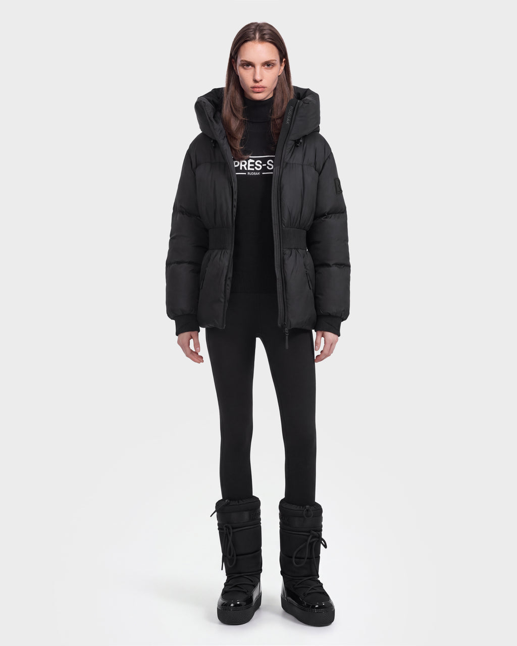 A person stands facing forward against a plain white background, dressed in the Rudsak DIAMOND - 8126772 Black puffer jacket with black leggings, black snow boots, and a black sweater.