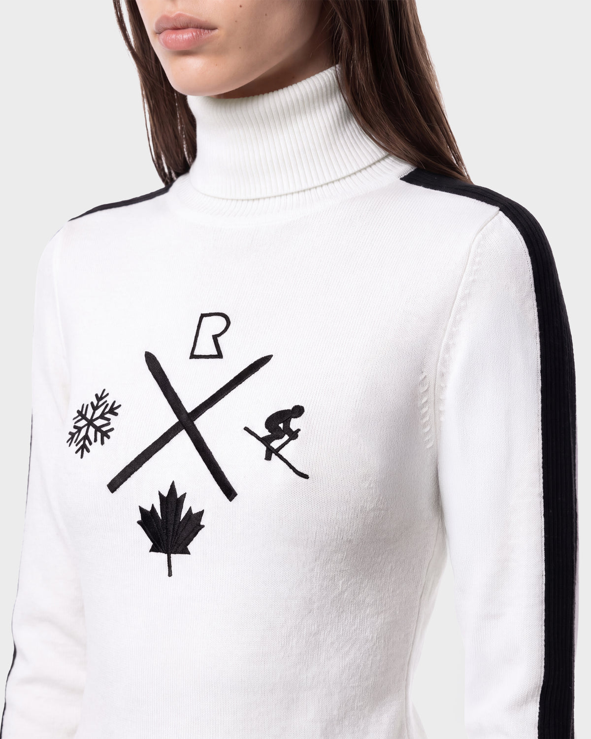 A woman makes a chic statement in her winter wardrobe, perfect for après-ski gatherings, wearing the Rudsak ELIZABETH - 8524623 Optic White women’s ski turtleneck—a white sweater featuring black shoulder stripes and black icons like a snowflake, skis, skier, leaf, and the letter R.