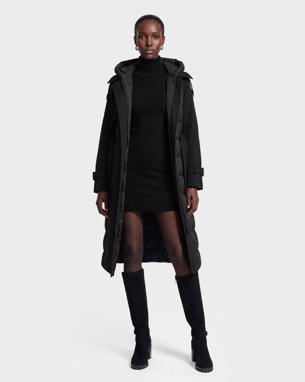 A woman stands facing forward, wearing the Rudsak ELVIE - 8126512 Black long puffer coat over a black turtleneck dress and knee-high black boots, against a plain white background.
