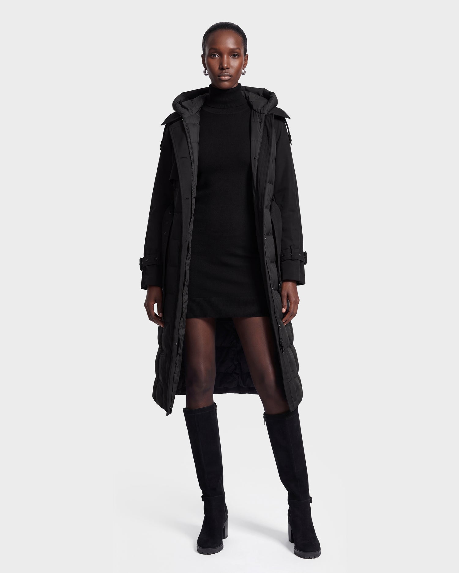 A woman stands facing forward, wearing the Rudsak ELVIE - 8126512 Black long puffer coat over a black turtleneck dress and knee-high black boots, against a plain white background.