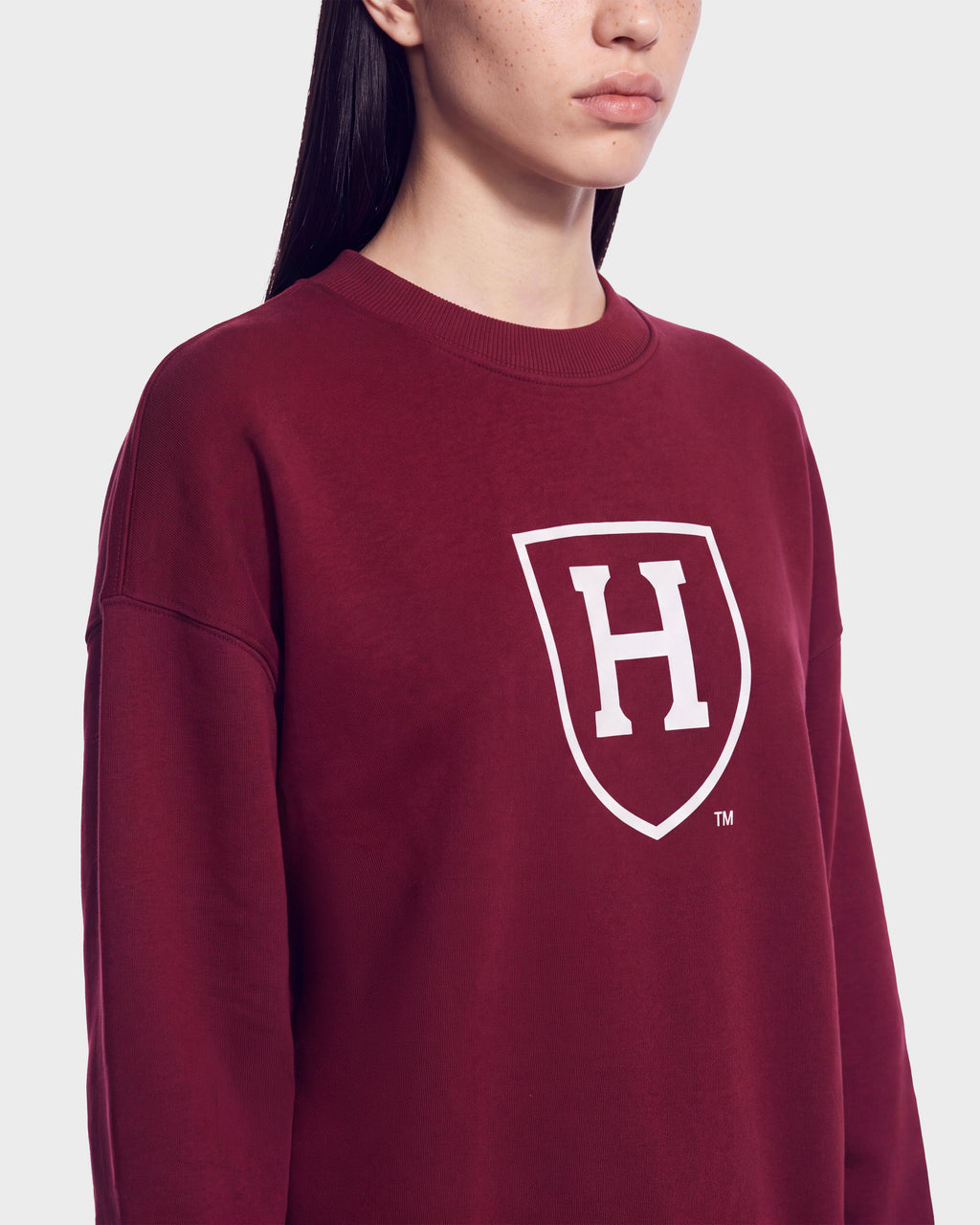 A person wearing the HARVARD ONE CRIMSON SWEATSHIRT 9524507 by Rudsak, a maroon unisex sweater with a white shield and prominent H logo on the front—an iconic piece inspired by classic Ivy League style—shown from the shoulders up against a plain background.