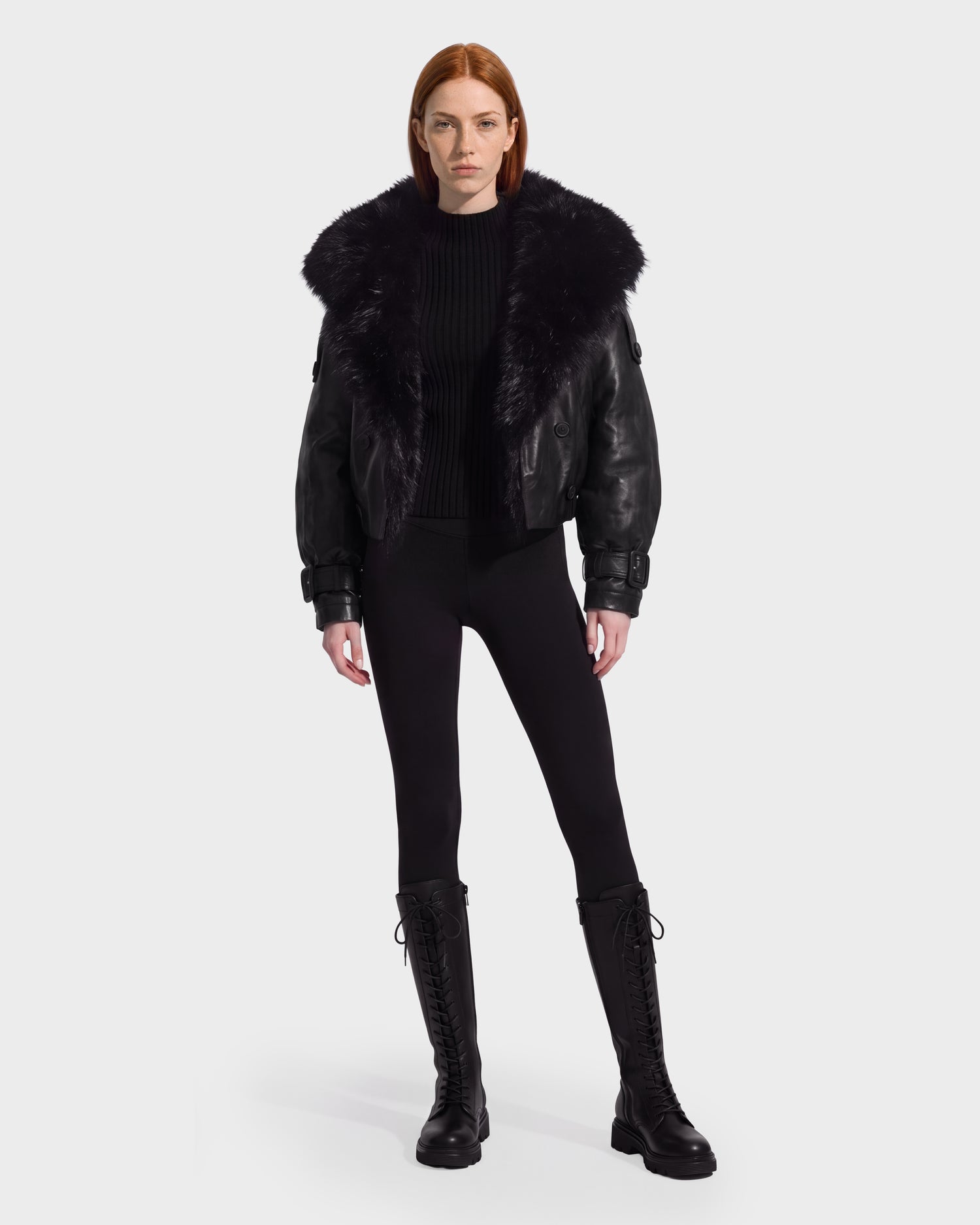A person with straight red hair wears the Rudsak JANELLE - 8126936 Black leather bomber featuring a faux fur collar, paired with a black sweater, black leggings, and tall black lace-up boots, standing against a plain light background for a modern silhouette.