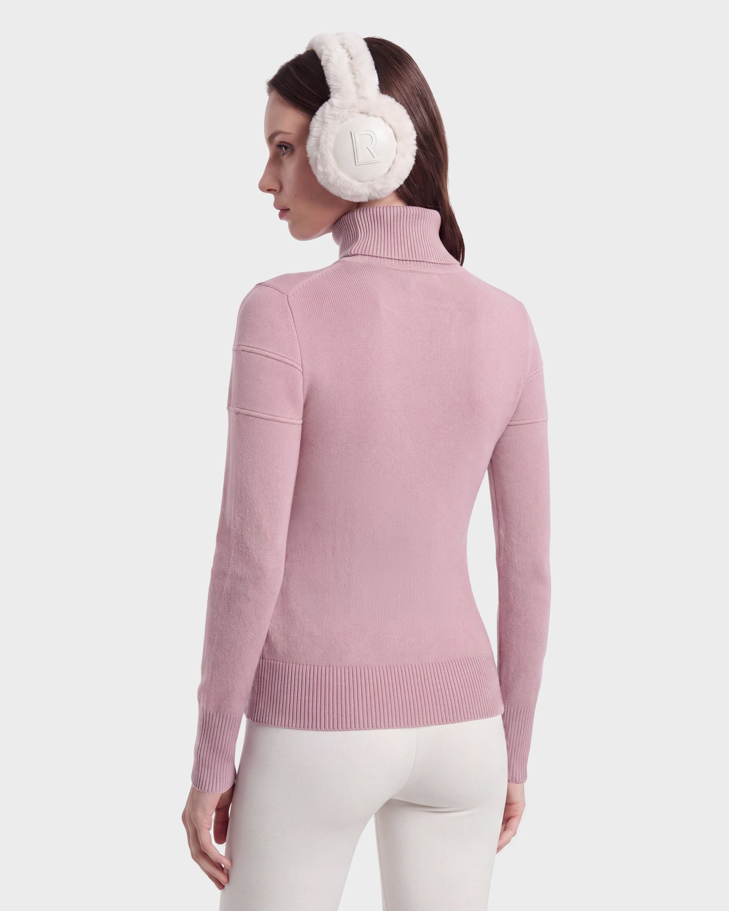 A woman facing away is wearing the ERIKA - 8525705 Vintage Rose turtleneck sweater by Rudsak, paired with white pants and white quilted earmuffs featuring the R logo, set against a plain light background.