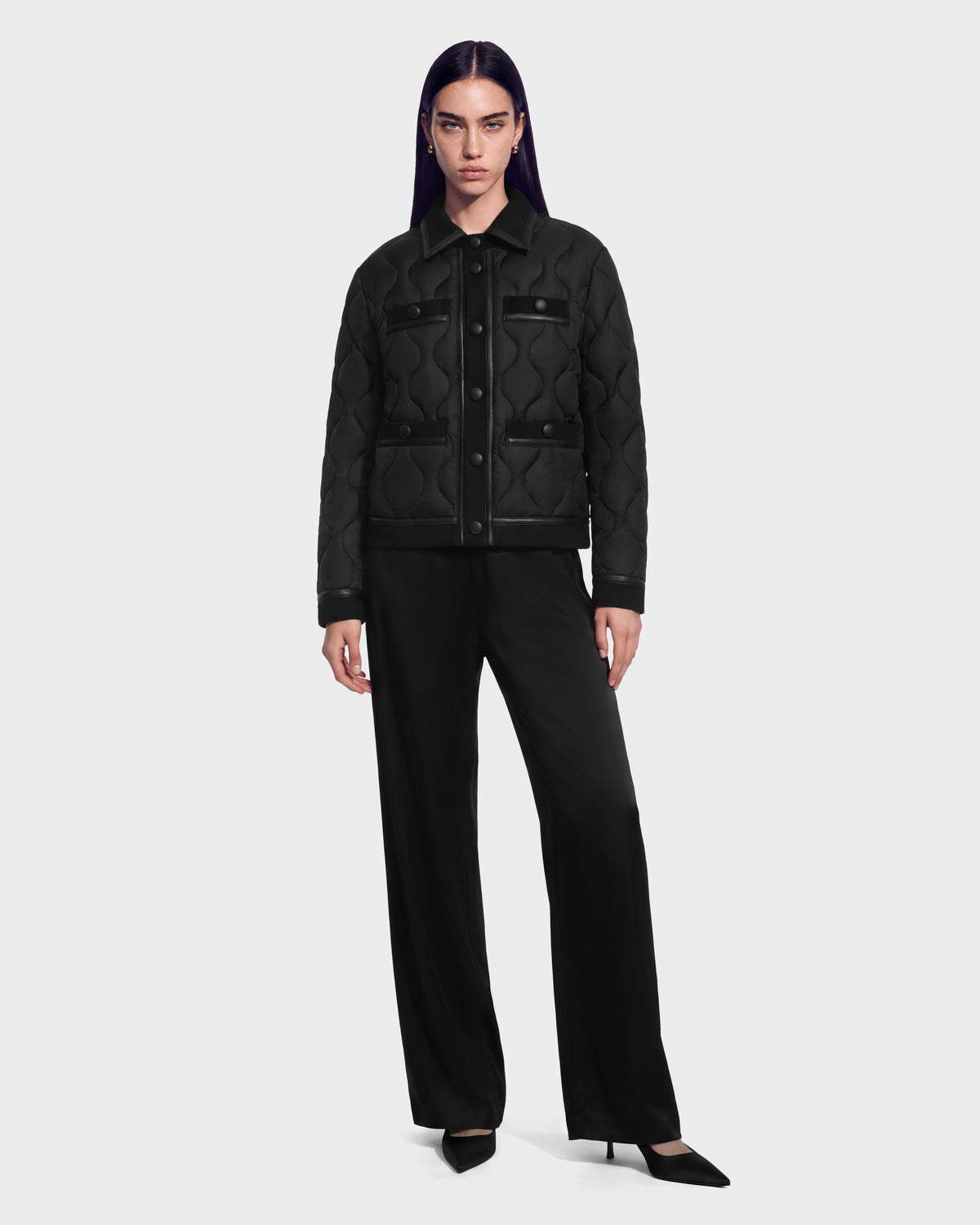 A person wearing a black JOLIE - 8125036 Black quilted down jacket with snap buttons and leather-trimmed pockets, paired with black trousers and pointed-toe heels.