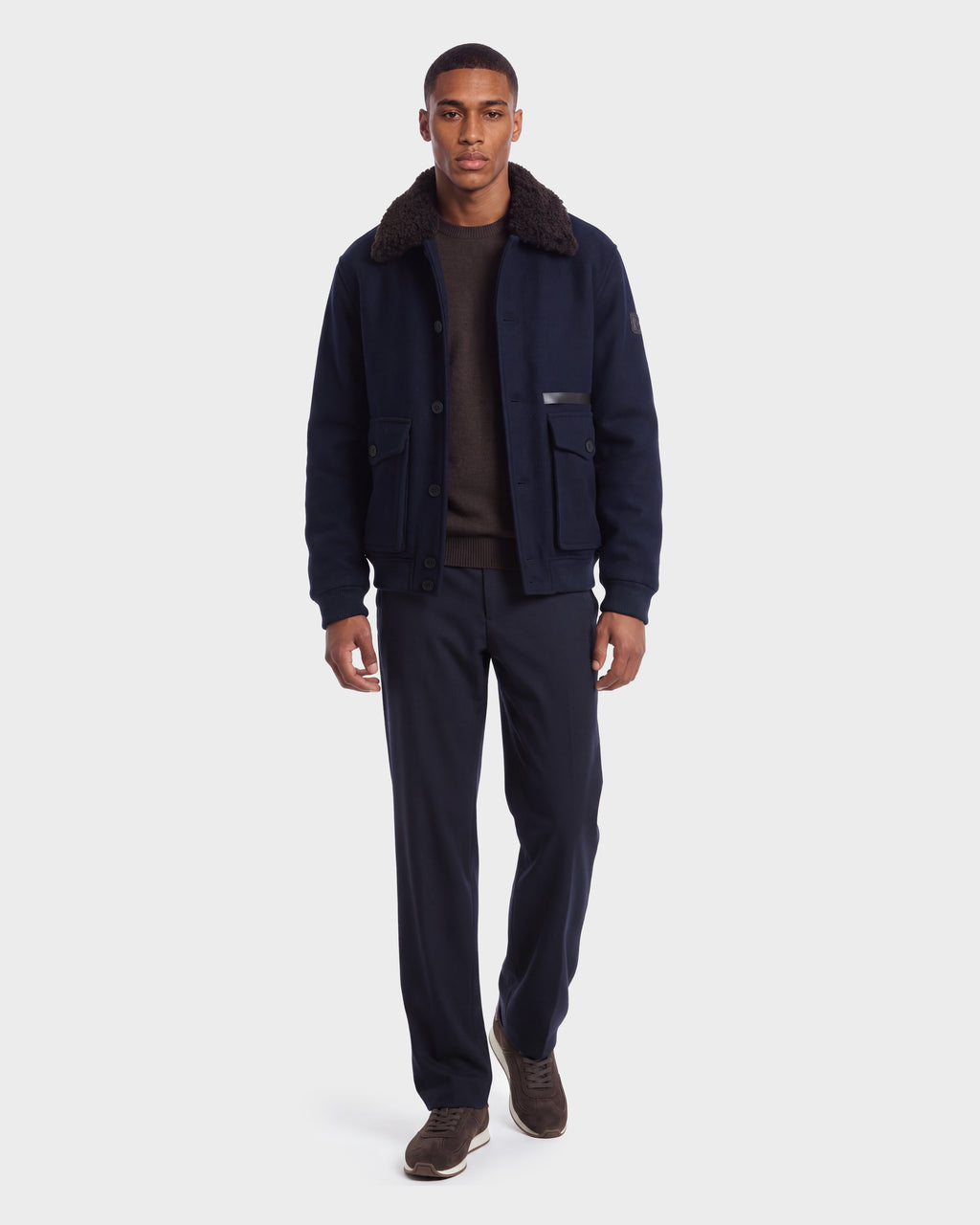 A man wearing a RUDSAK JULIUS - 6126723 Navy bomber jacket with a dark faux-fur collar over a brown sweater and navy trousers.