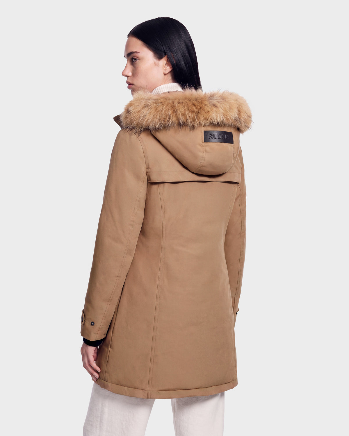 A woman with dark hair stands sideways wearing the Rudsak KEYSHIA - 8126517 Camel down parka with a fur-lined hood, styled with light pants and a turtleneck.