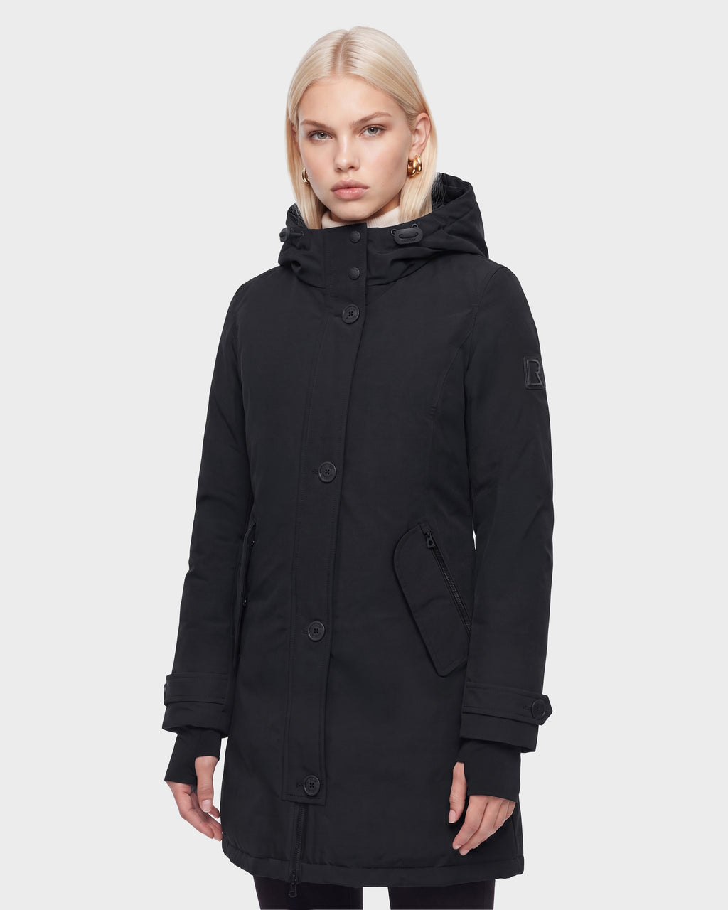 A woman with blonde hair wears the Rudsak KEYSHIA - 8126517 Black long down winter coat, featuring large buttons, side pockets, and a high collar, against a plain white background.