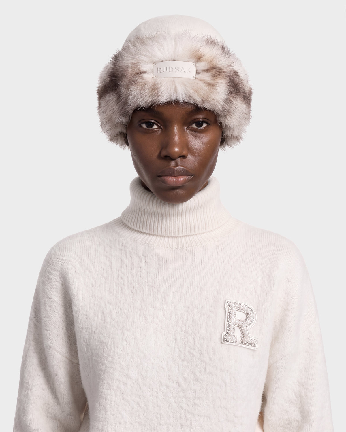 A person wearing a LORETTA W - 8426510 Vanilla cream-colored turtleneck sweater with an embroidered R patch and a RUDSAK faux fur hat featuring a front logo patch.