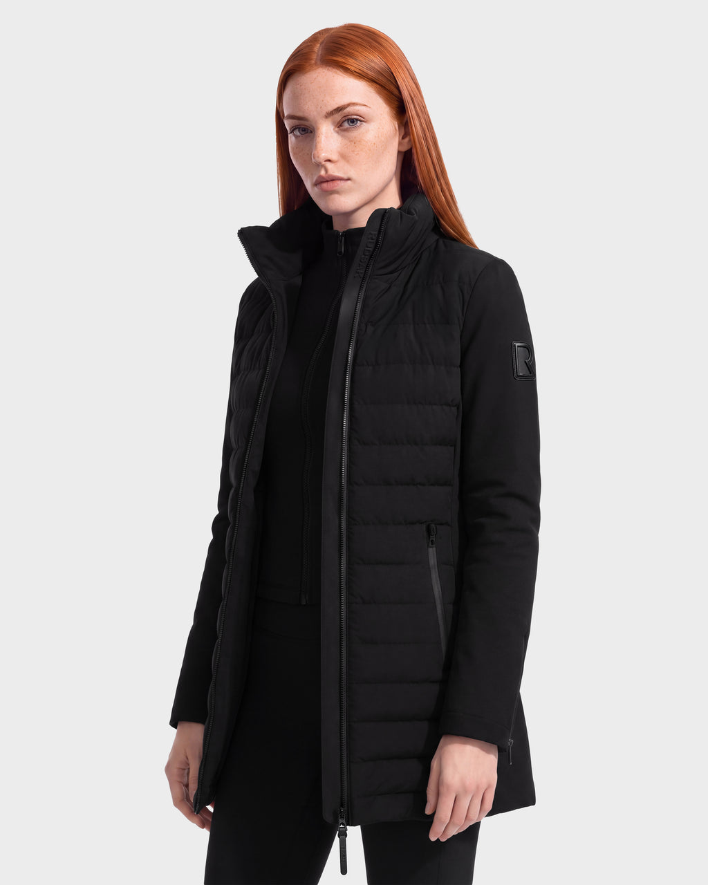 A woman with long red hair wears the Rudsak MERINA - 8125056 Black jacket, a black quilted down jacket featuring a high collar and zippered pockets, standing against a plain light background. This women’s jacket from Rudsak is the perfect stylish and practical lightweight transitional layer for changing seasons.