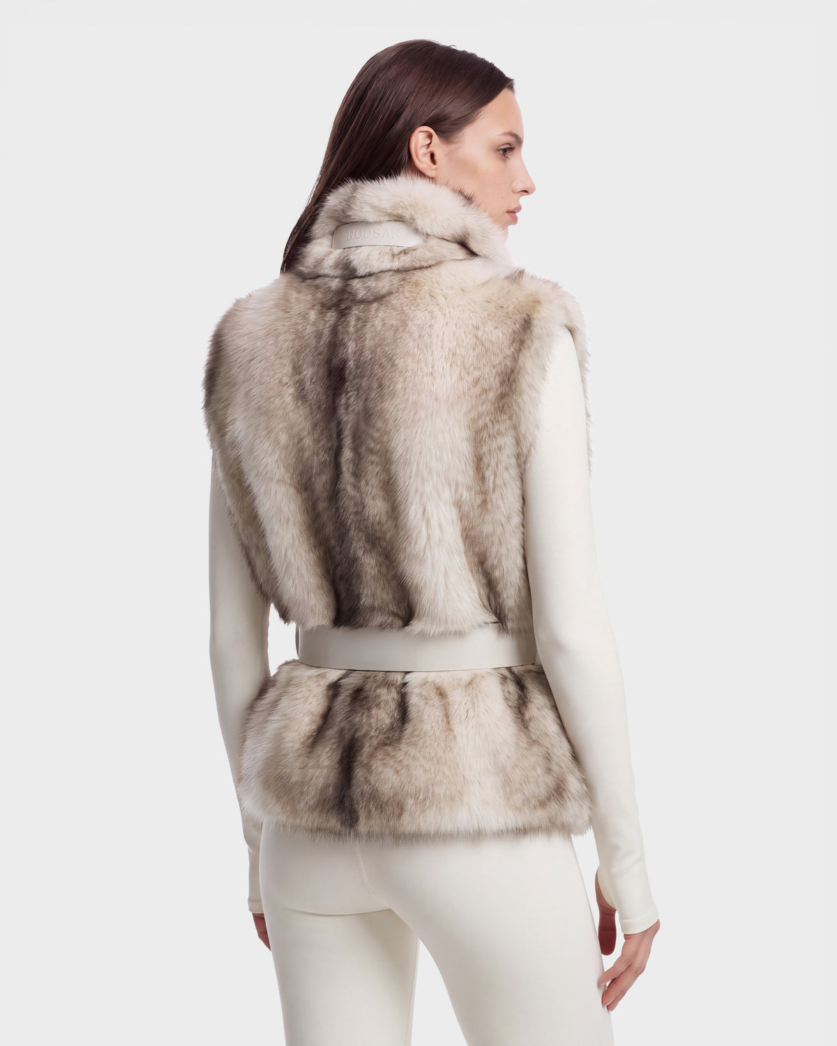 A person viewed from behind wearing a white mock-neck pullover and a Rudsak MAULIN - 8524611 Mix Vanilla belted, waist-length faux fur vest in shades of cream and grey.
