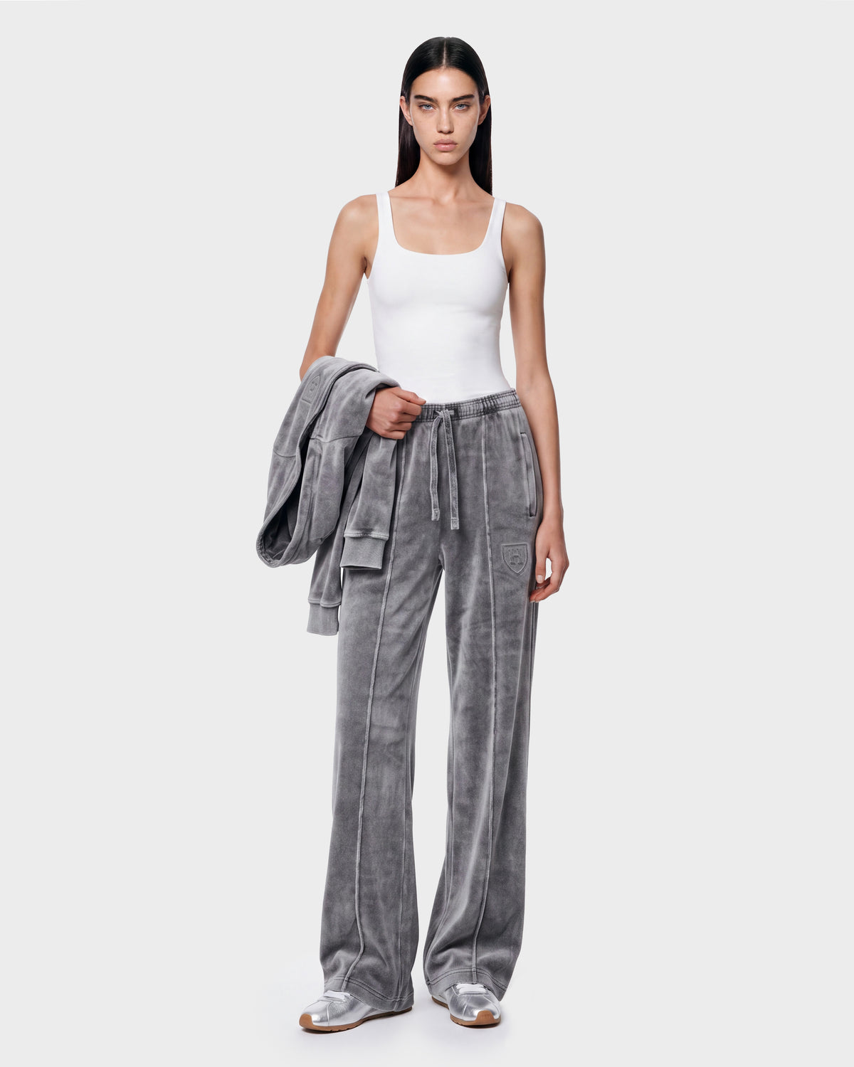 A person with long dark hair stands against a plain light background wearing loose, gray velour OLIVYA HV - H526903 Titanium pants by Rudsak and a white tank top, while holding a matching gray jacket.