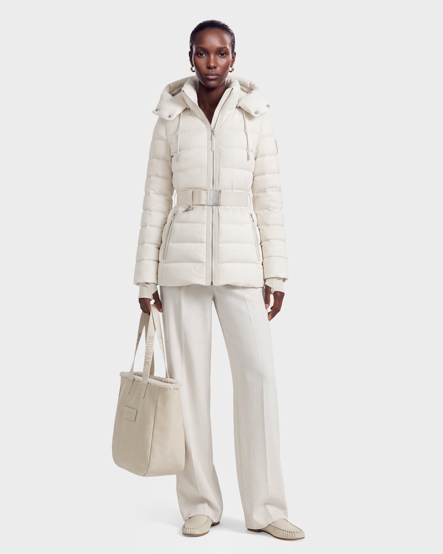 A woman wearing the Rudsak PELIN S - 8124813 Vanilla belted, hooded down puffer jacket, paired with cream wide-leg pants and loafers, holds a large cream tote bag. She stands facing forward against a plain light background.