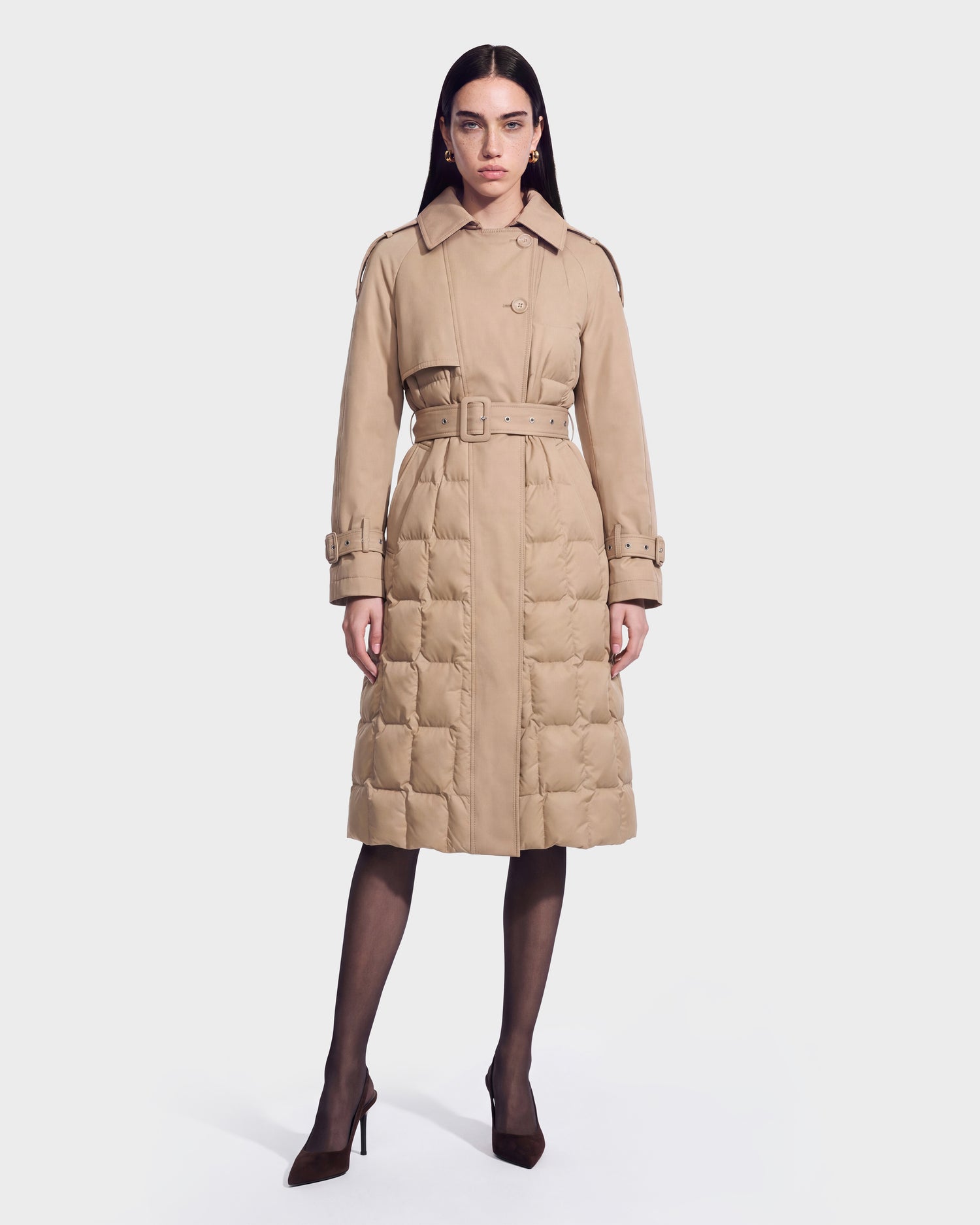 A woman stands against a plain white background wearing the Rudsak ELVIE - 8126512 Almond, a long, beige belted down trench coat with a quilted lower half. She pairs it with black sheer tights and brown high heels.