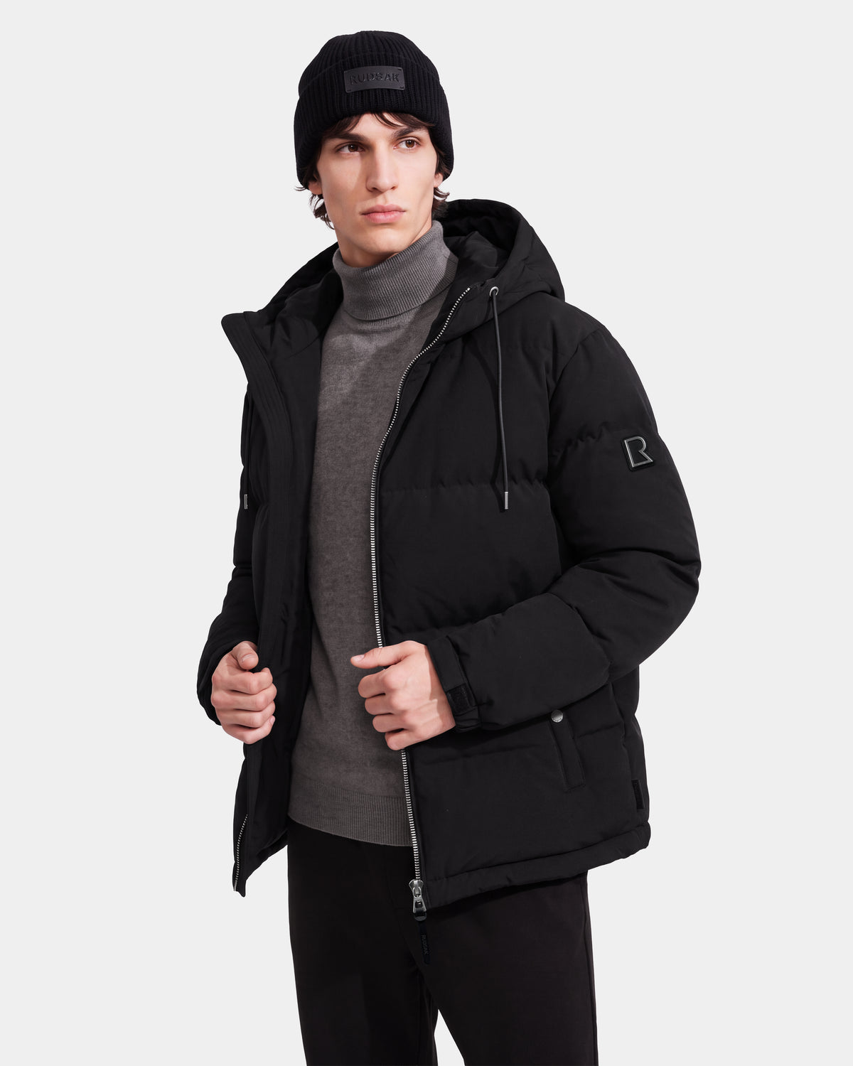 A man wears the STERLING EL - 6126759 Black winter jacket by Rudsak, paired with a gray turtleneck and black beanie, standing against a plain light background with one hand holding the jacket’s lapel and the other in his pocket.