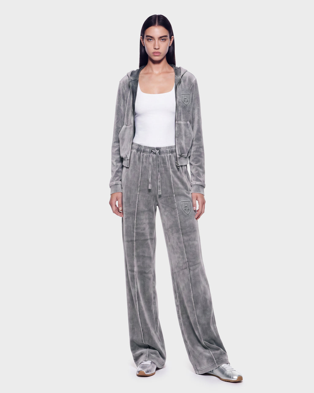 A person is wearing the Rudsak TYRA HV - H526904 Titanium velour tracksuit, which features a hooded jacket, paired with a white tank top and silver sneakers. They stand against a plain light background, facing forward with their arms at their sides.