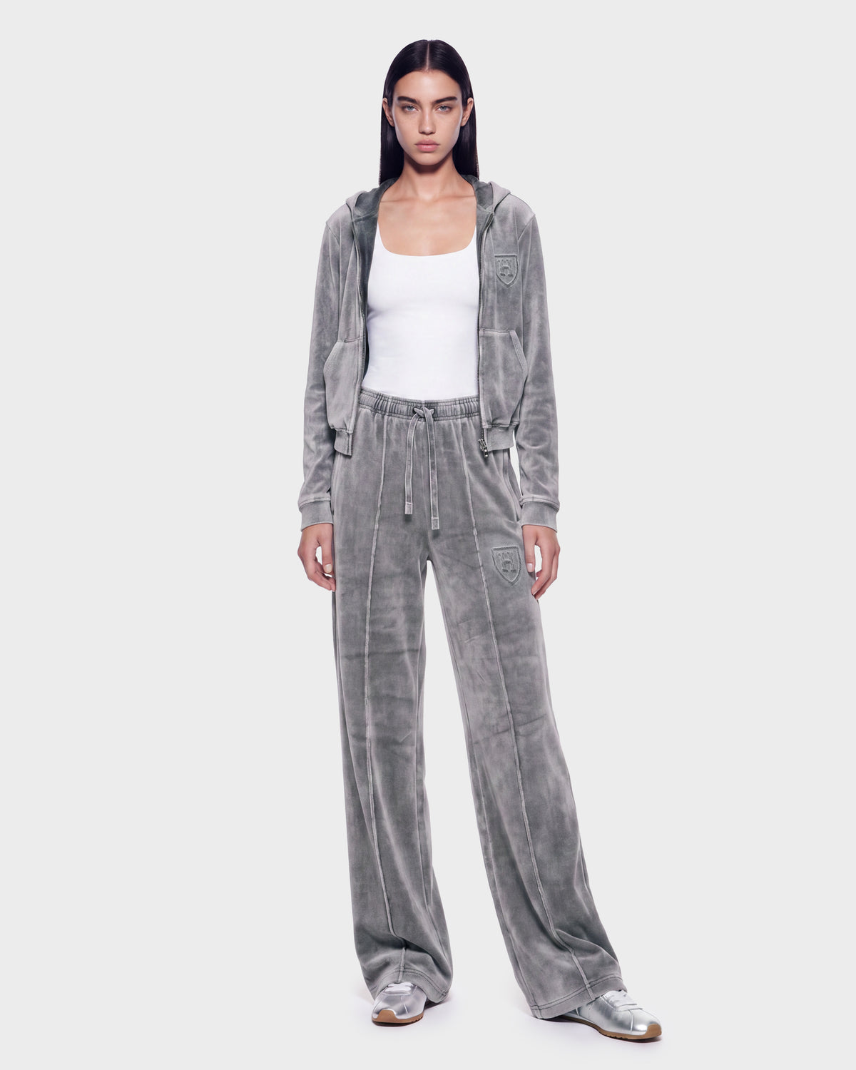 A person is wearing the Rudsak TYRA HV - H526904 Titanium velour tracksuit, which features a hooded jacket, paired with a white tank top and silver sneakers. They stand against a plain light background, facing forward with their arms at their sides.