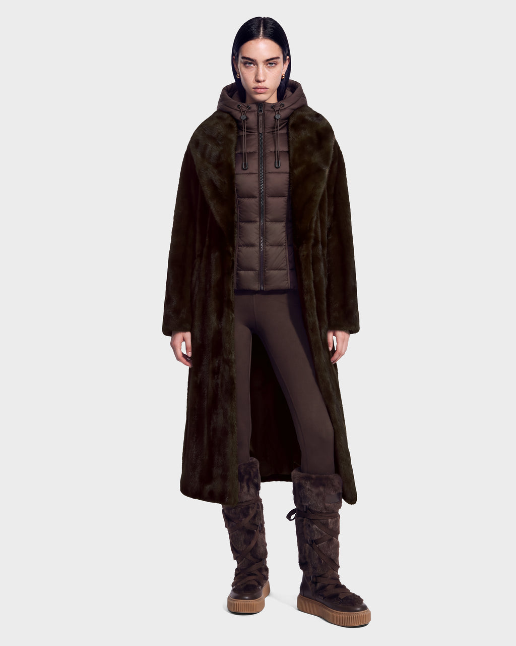 A model wears a VIVIENNE - 8126528 Espresso long, dark faux-fur coat over a brown quilted duck down jacket, leggings, and tall, fur-lined boots with thick rubber soles, capturing the essence of winter city dressing.