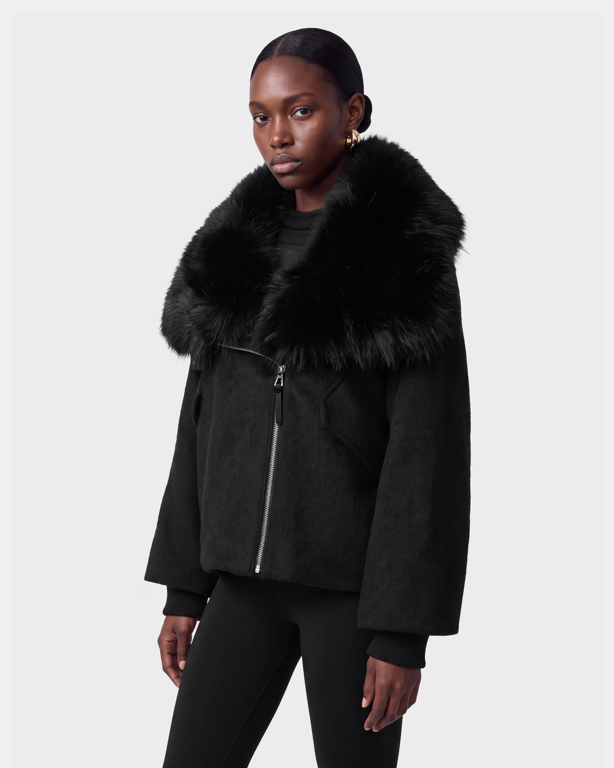 A woman models the Rudsak ZARIA - 8126999 Black jacket, featuring a faux fur collar, paired with black pants against a plain light gray background. She gazes slightly to the side with a neutral expression.