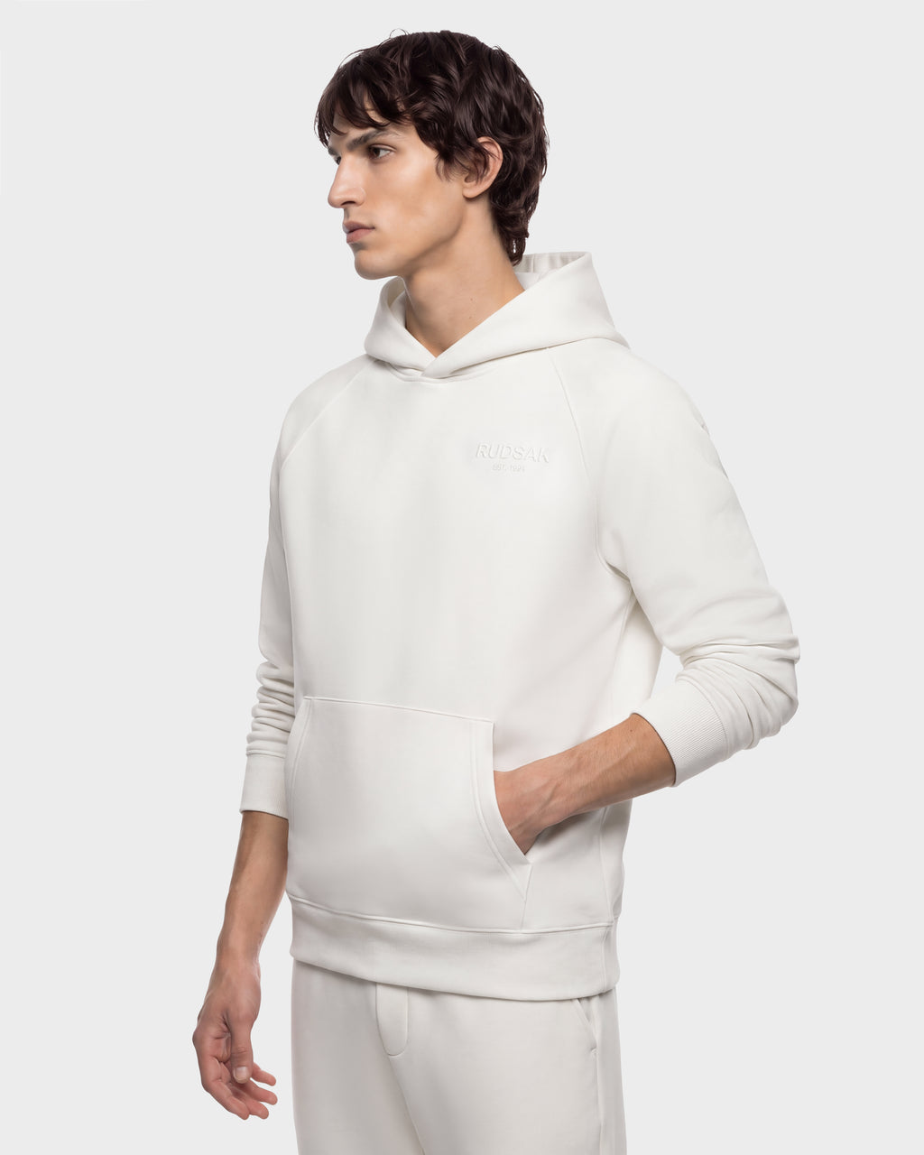 A man wears a Rudsak BRIAR - 9525001 Vanilla M, a premium cotton blend, cream-colored pullover hoodie with a front kangaroo pocket, creating a casual look.