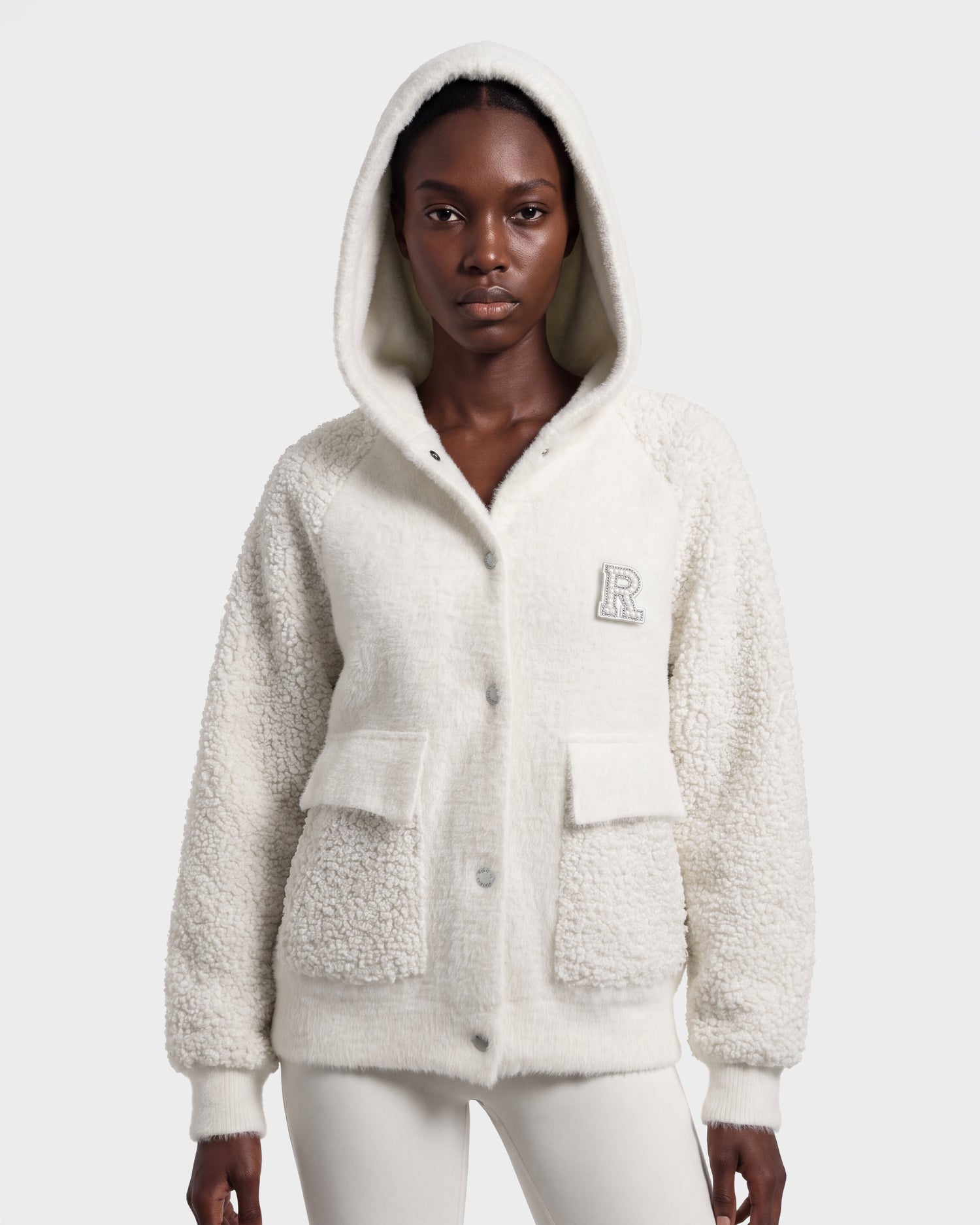 A person wearing the DAKOTA - 8526515 Iceland cream-colored hooded fleece jacket by Rudsak, featuring large front pockets and an R patch on the chest, stands against a plain background.