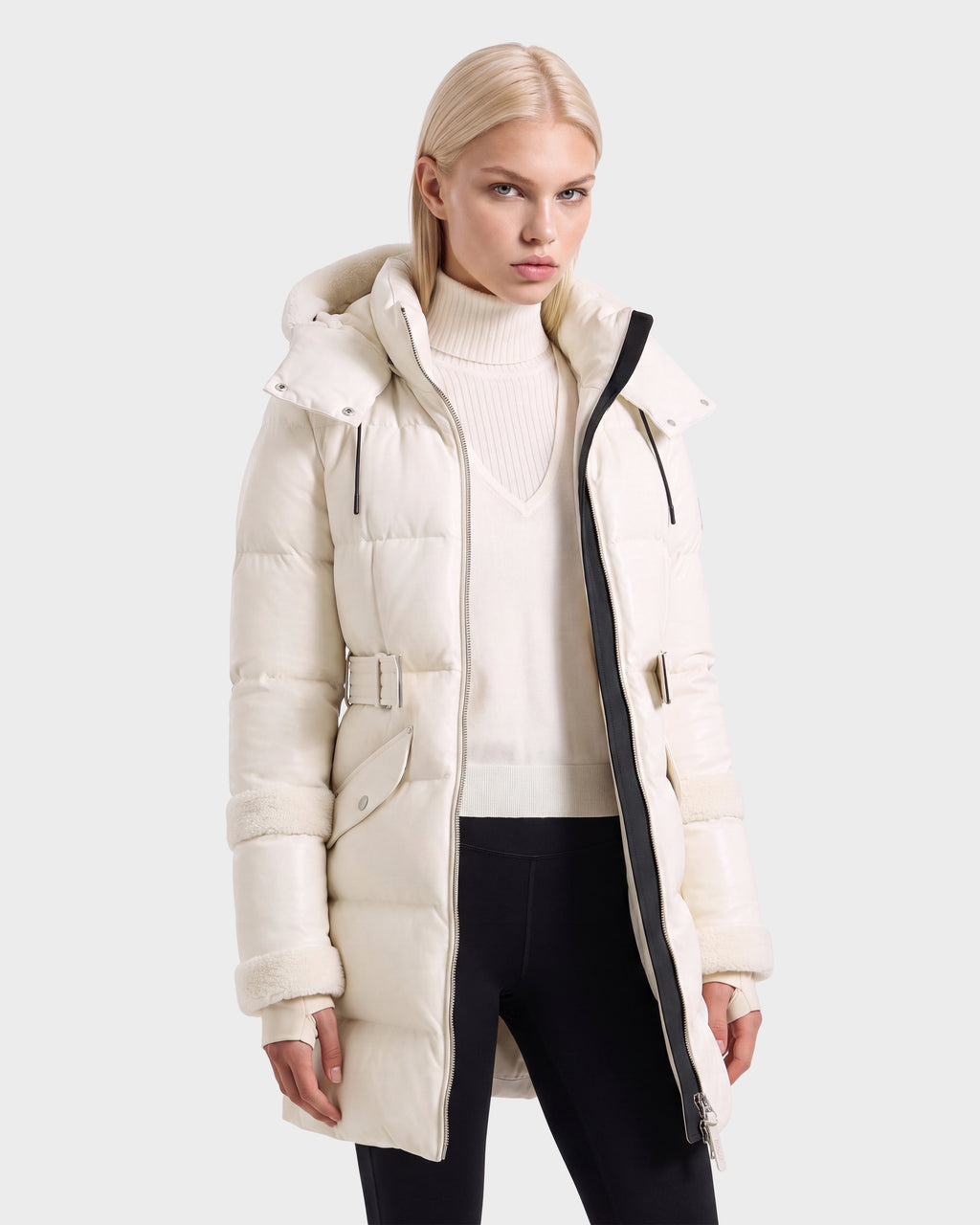 Woman wearing a cream-colored Rudsak AUDREY L - 8124585 Vanilla knee-length down puffer jacket with a hood and belted waist over a matching turtleneck sweater and black leggings.