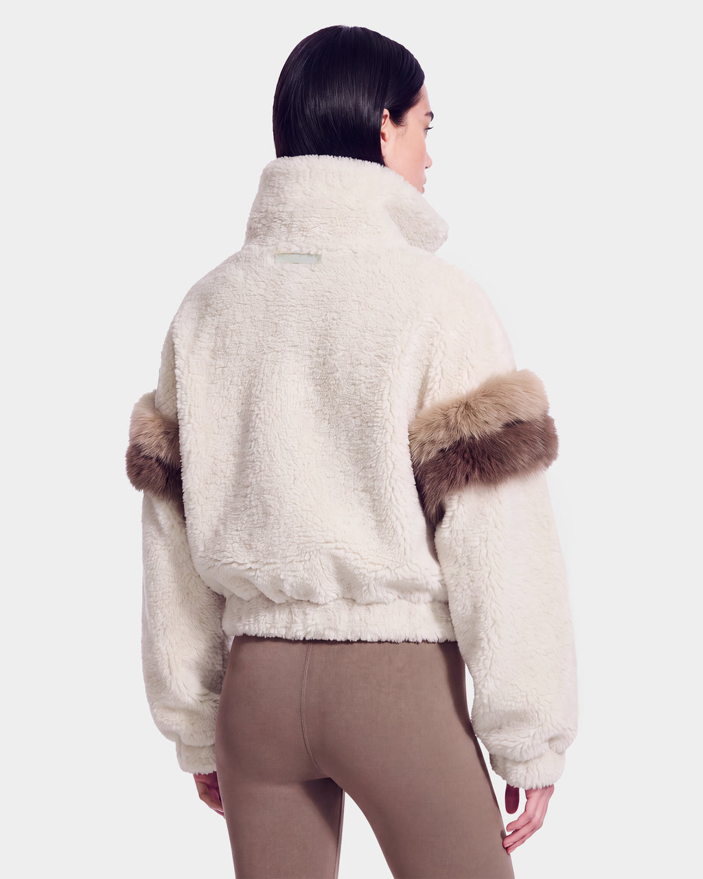 A person shown from the back against a plain light background is wearing the Rudsak ALEIA - 8526693 Vanilla jacket, a white, fluffy piece featuring brown fur trim on the sleeves, paired with taupe leggings.