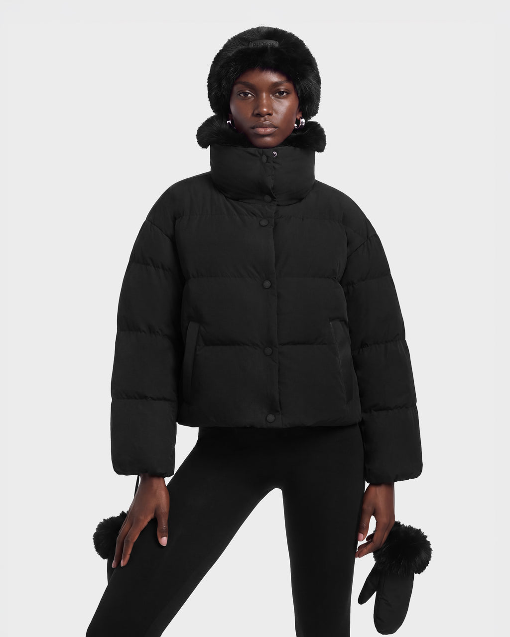 A model wears the ALONDRA - 8126992 Black cropped puffer jacket with a high collar and fur-trimmed hood, paired with matching black leggings, a fur-trimmed hat, and fur-accented mittens.