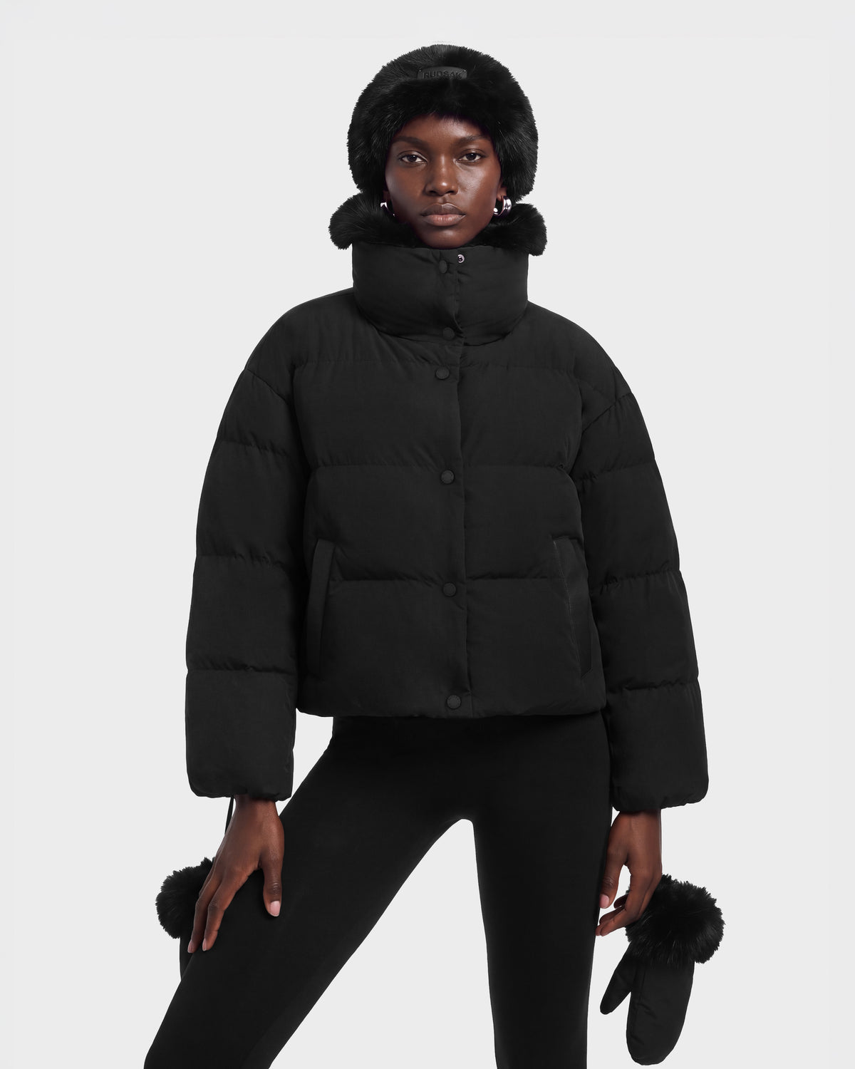 A model wears the ALONDRA - 8126992 Black cropped puffer jacket with a high collar and fur-trimmed hood, paired with matching black leggings, a fur-trimmed hat, and fur-accented mittens.
