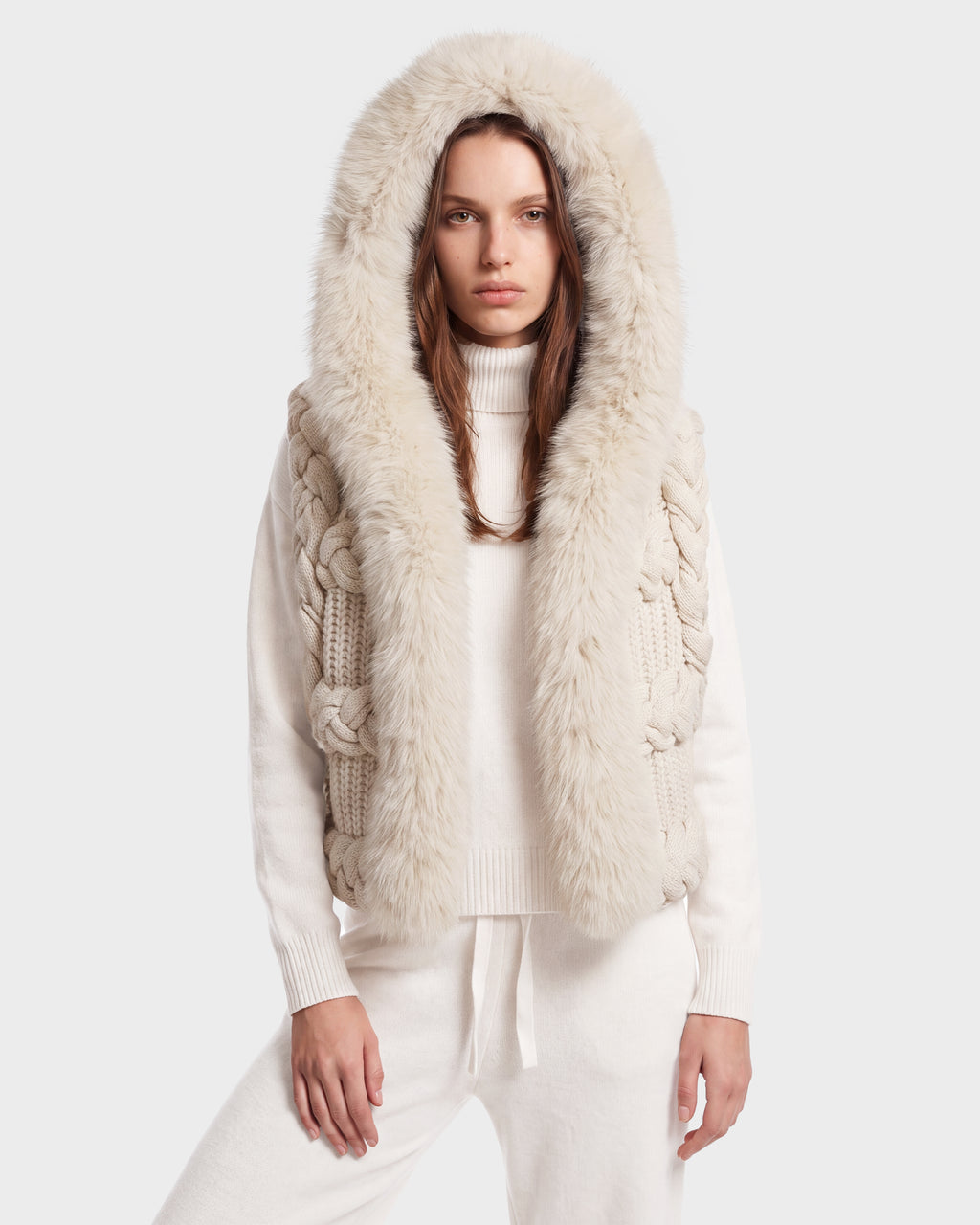 A woman stands facing forward against a plain background, effortlessly showcasing winter layering in the CICELY - 8526686 Vanilla sleeveless knit vest by Rudsak, featuring a cozy cream-colored design with a large, fluffy faux fur-trimmed hood.