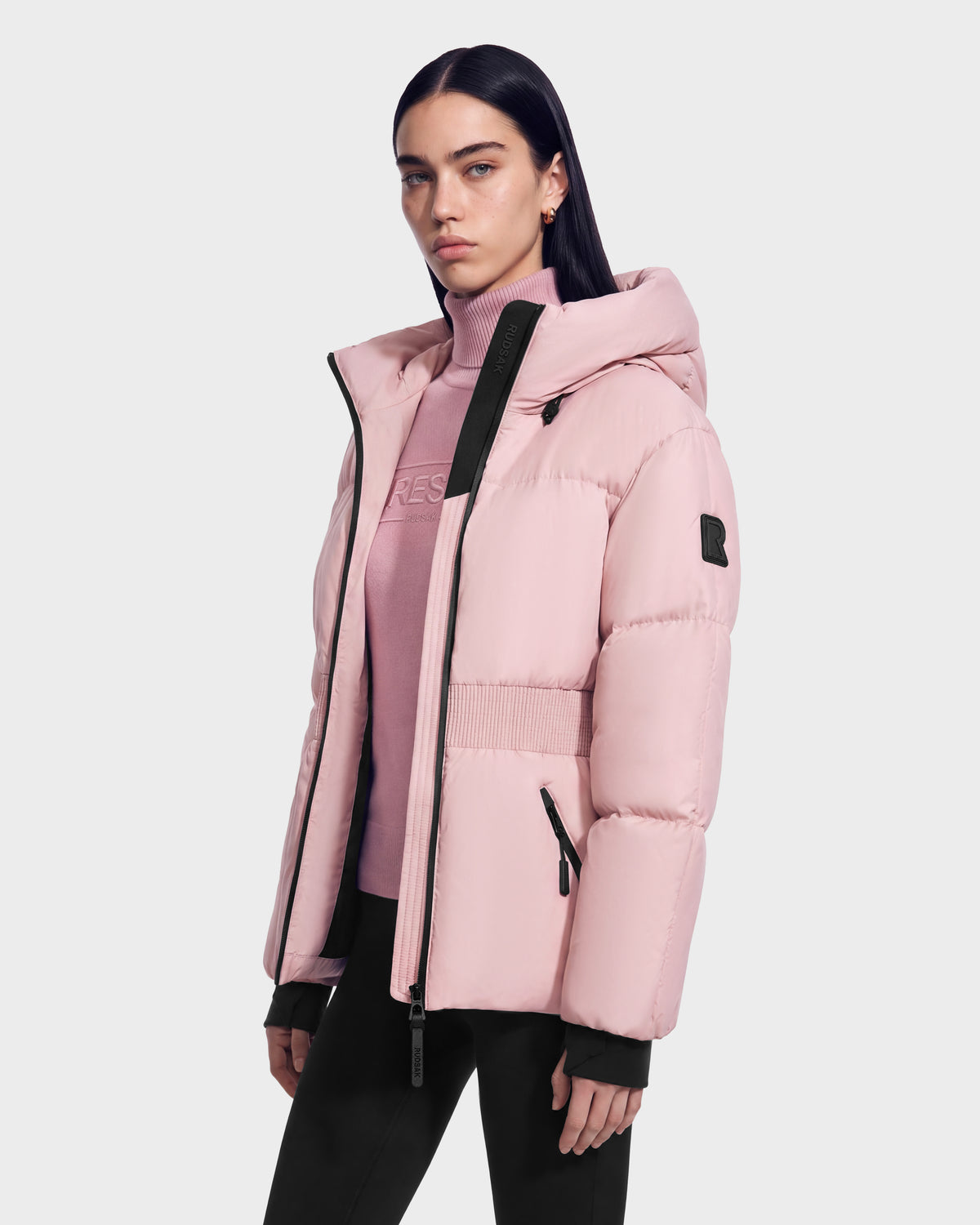 A woman wearing a Rudsak DIAMOND - 8126772 Vintage Rose pale pink quilted puffer jacket with black zipper details over a matching pink turtleneck sweater for winter warmth.