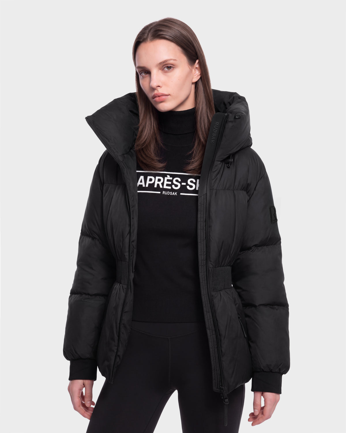 A woman wears the Rudsak DIAMOND - 8126772 Black puffer jacket over a black APRÈS-SKI sweater, standing against a light gray background and looking at the camera with a neutral expression.