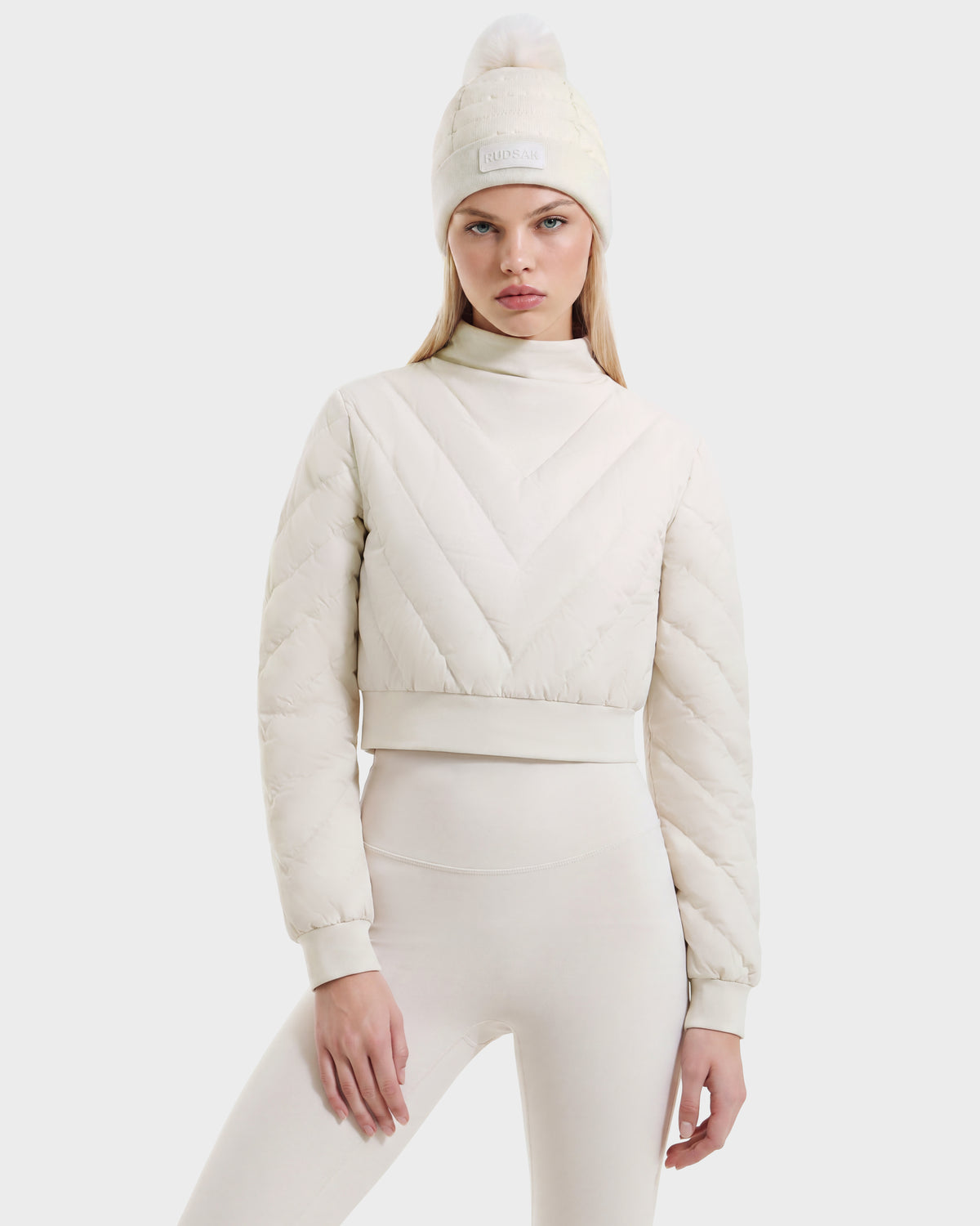 A person wears a cream-colored KATYA - 8526531 Vanilla quilted mock-neck cropped down sweatshirt and a matching knit beanie with a pom-pom.
