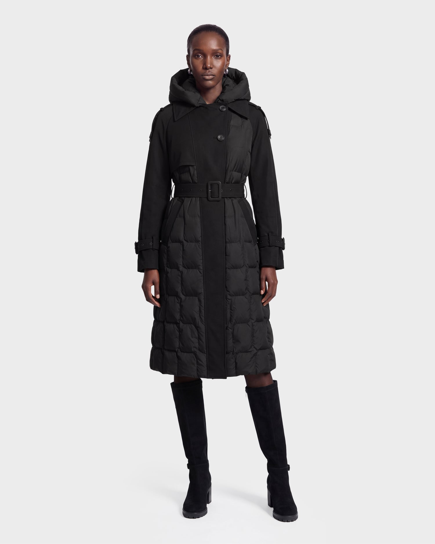 A person stands facing forward against a plain light background, wearing the Rudsak ELVIE - 8126512 Black long belted hooded coat paired with tall black boots.