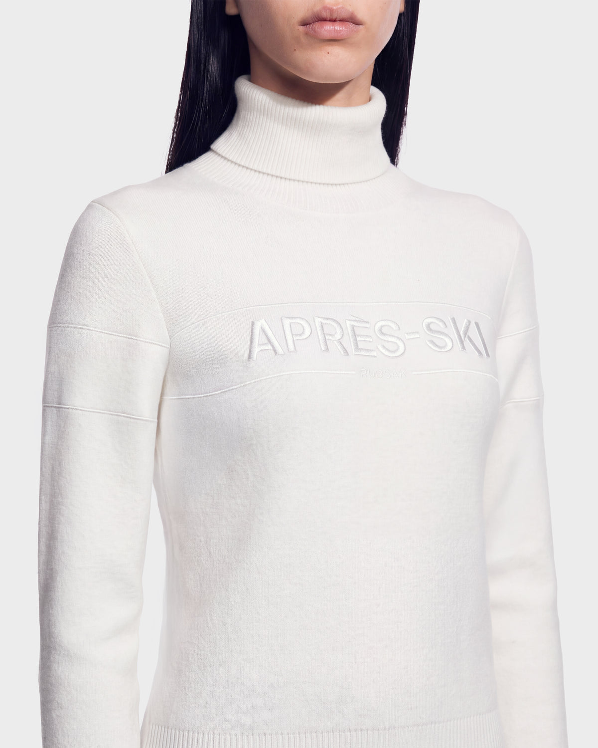 A woman wearing the ERIKA - 8525705 Iceland sweater by Rudsak, a cozy white turtleneck embroidered with “APRES-SKI” across the chest. This essential winter wardrobe piece is showcased against a plain, light-colored background.