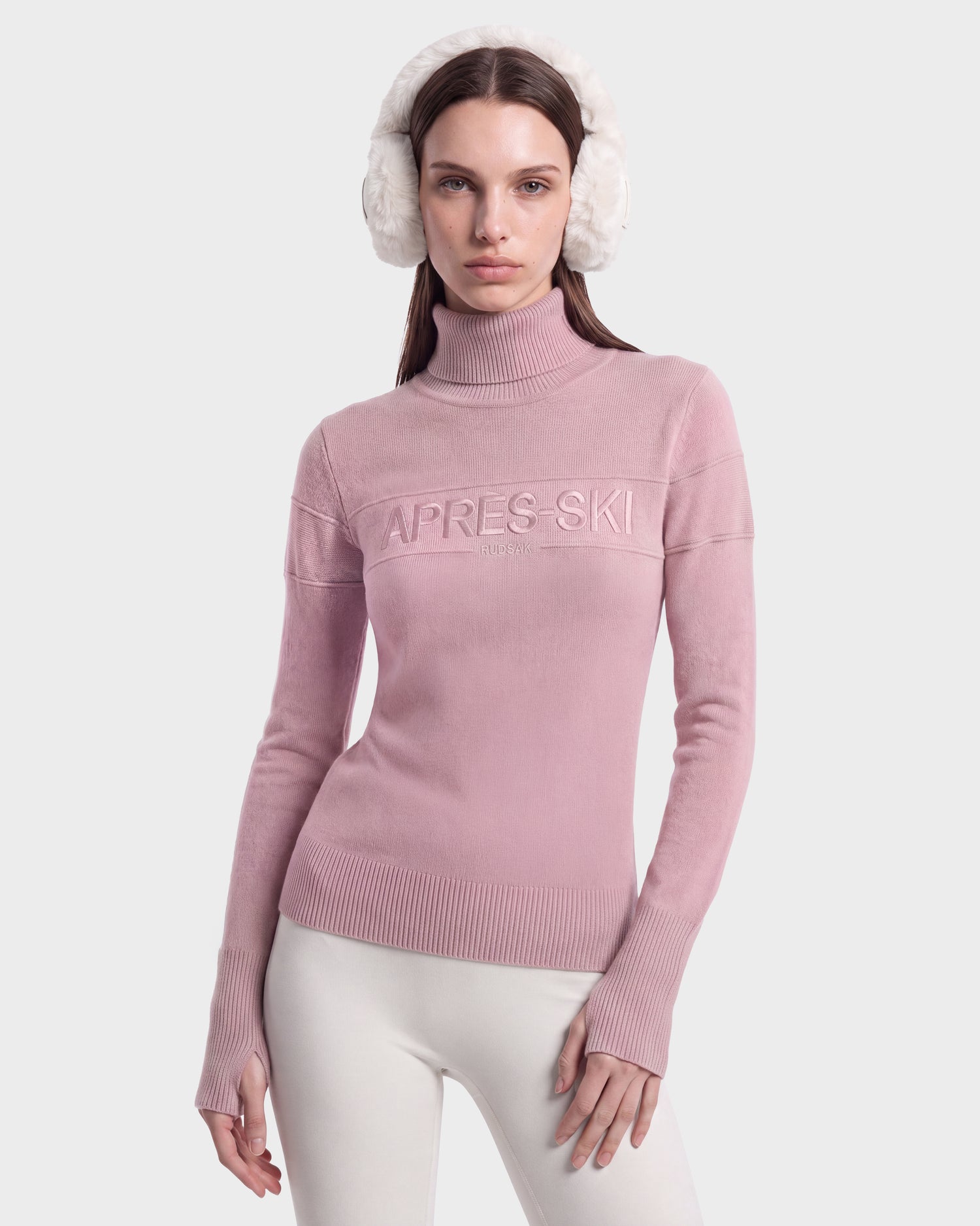 A woman wearing white earmuffs and the Rudsak ERIKA - 8525705 Vintage Rose apres-ski sweater, featuring “APRES SKI” text and paired with white pants, poses against a plain light background, highlighting this chic addition to your winter wardrobe.